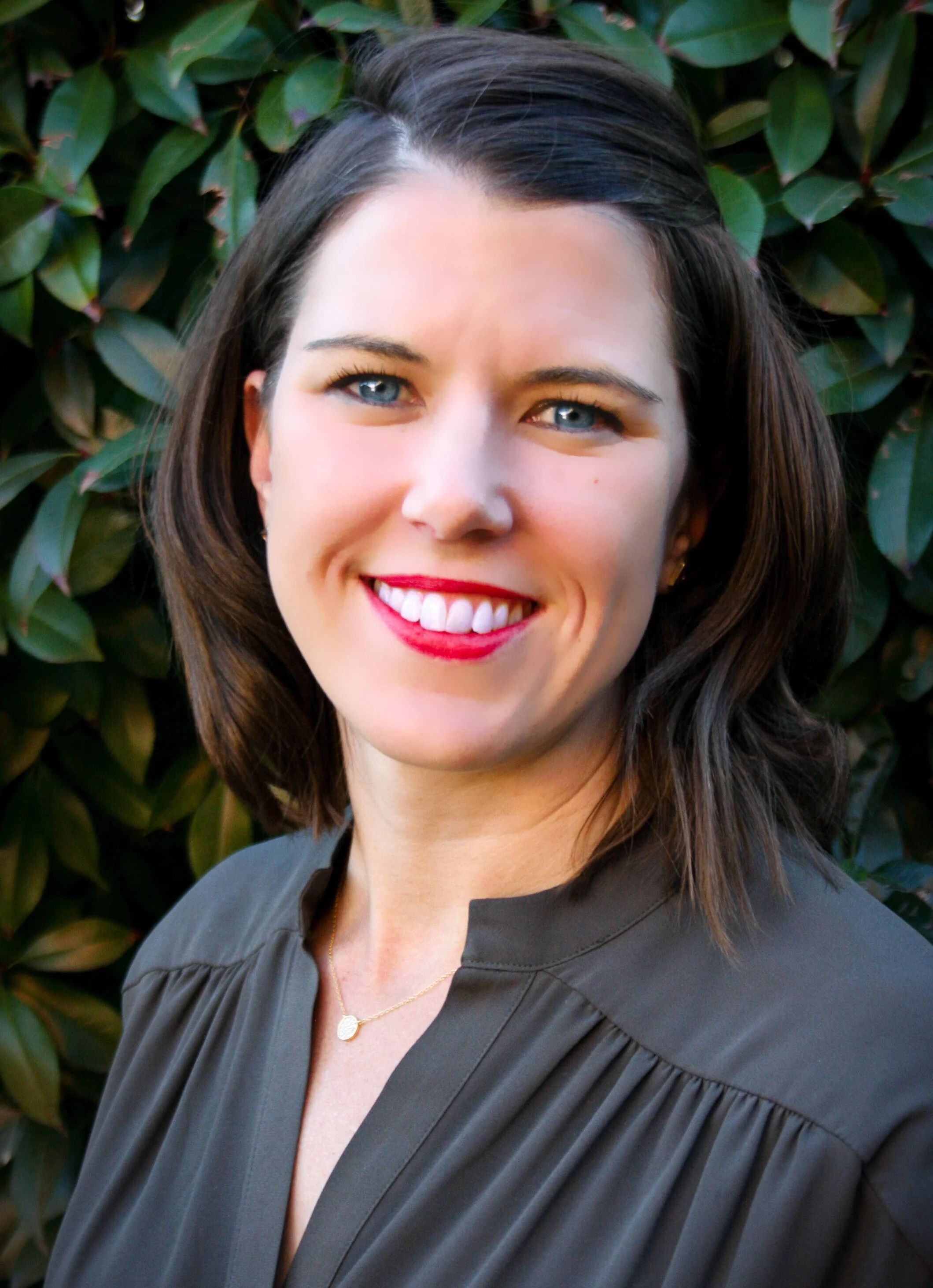 A woman with brown hair and bright blue eyes smiling, wearing red lipstick, a gray top, and a gold necklace, standing outdoors in front of green foliage.