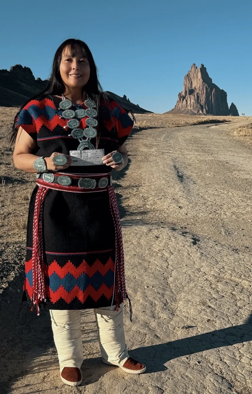 A woman standing on a dirt path in a desert landscape with a large rock formation in the background, wearing traditional Native American clothing and jewelry.