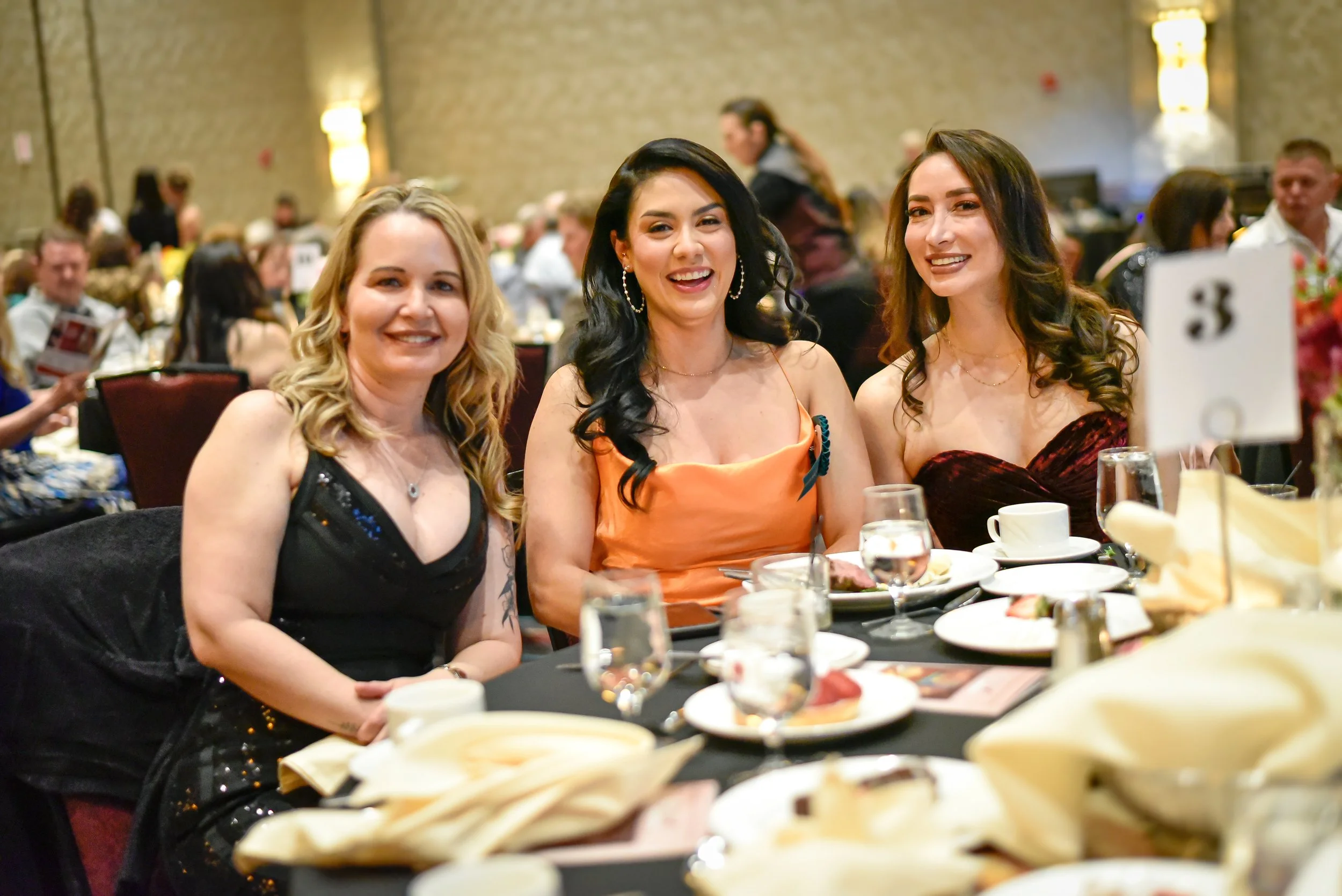 Three women sitting at a banquet table, smiling at the camera, in a large decorated event hall filled with other guests.