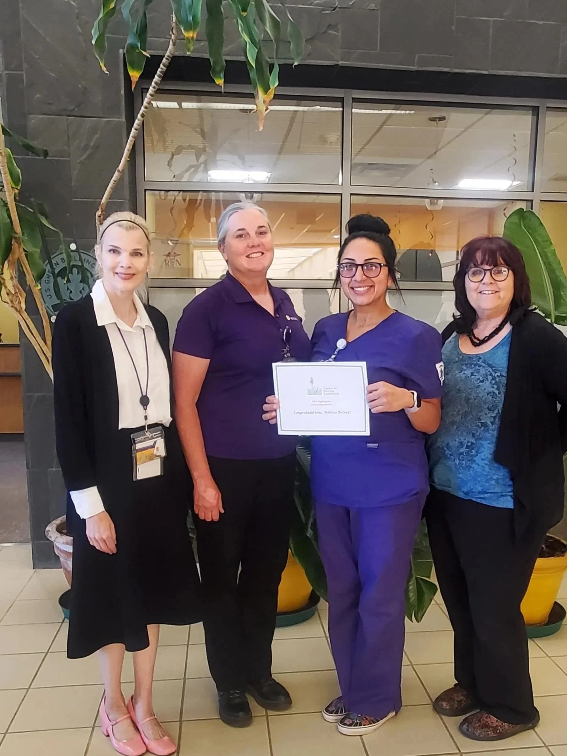 Four women standing together indoors, with the woman second from the right holding a certificate of recognition, smiling for the photo.
