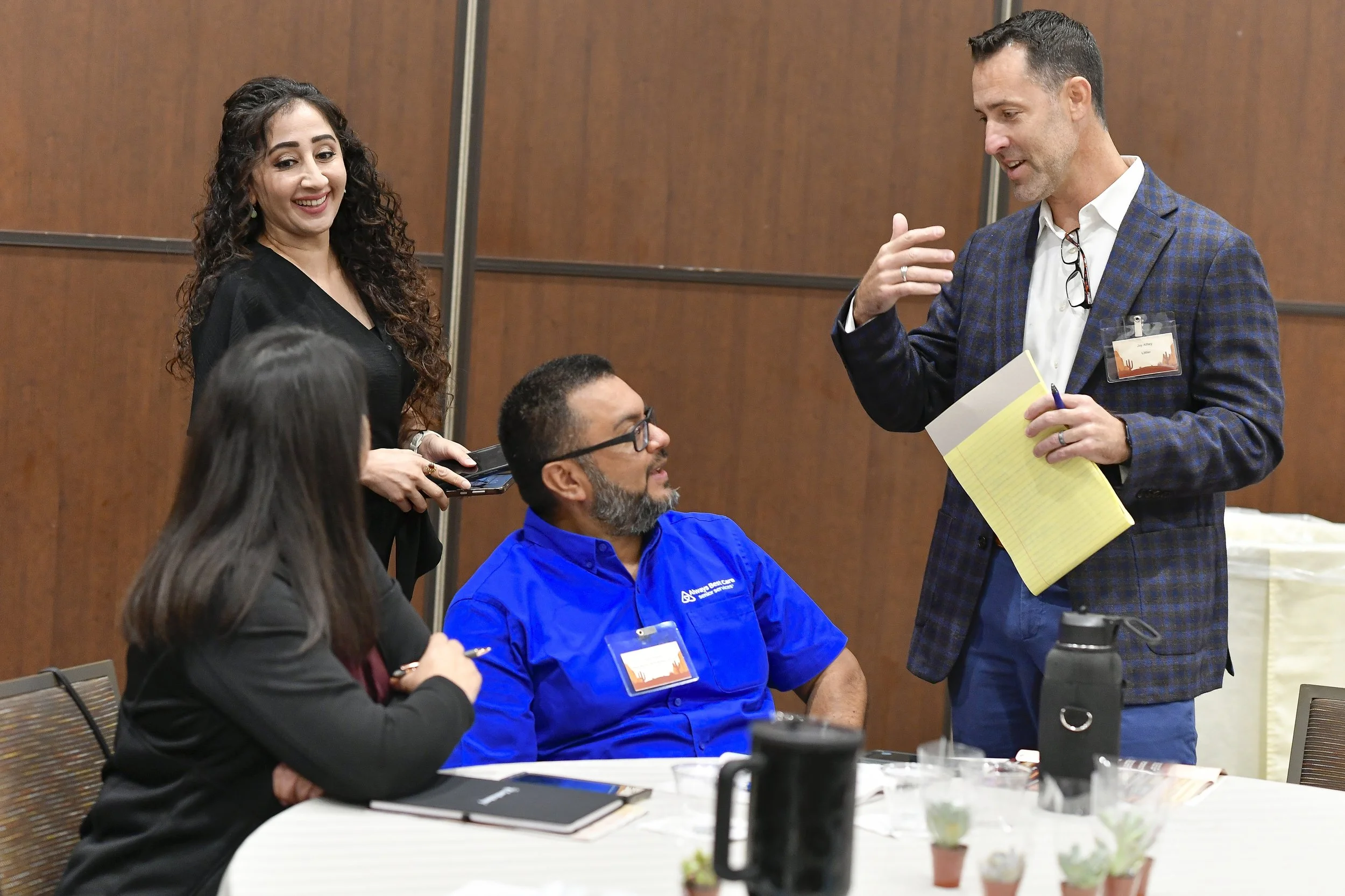 Group of four diverse professionals in a meeting room, engaging in conversation, with one man standing and speaking, and the others seated around a table.