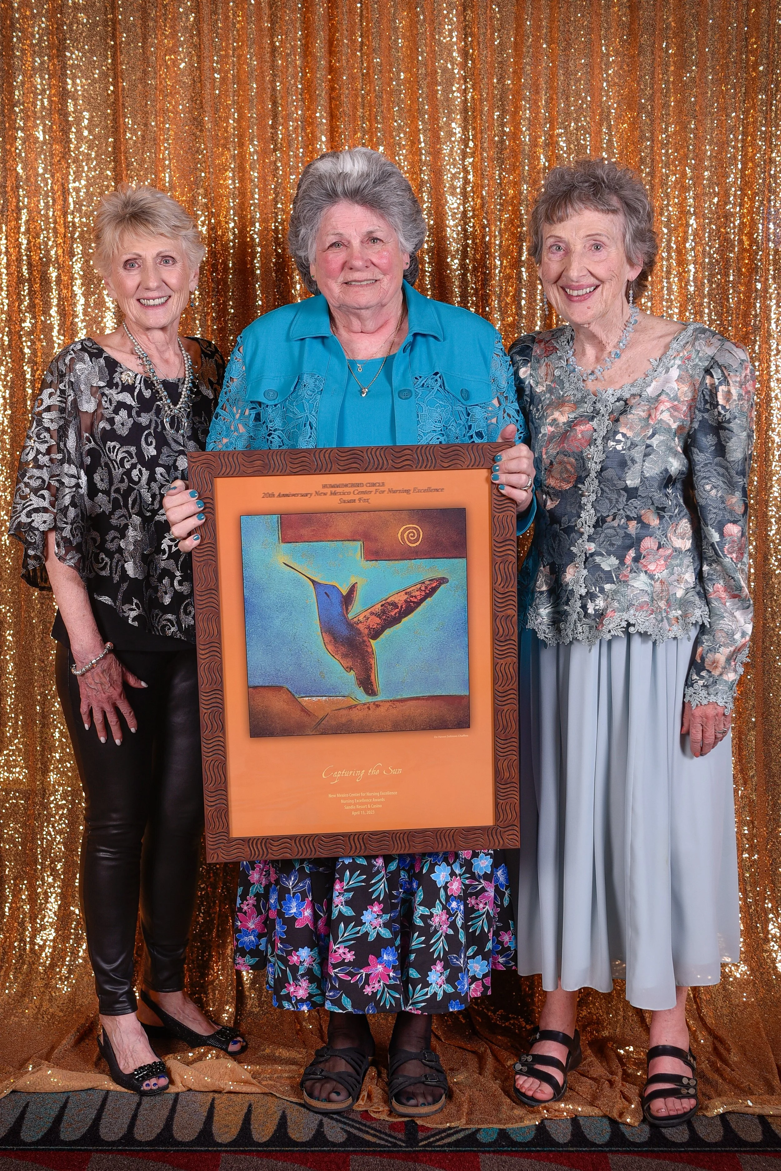 Three elderly women standing together in front of a gold sequin backdrop, holding a framed artwork of a hummingbird. They are dressed in colorful, patterned clothing and are smiling.
