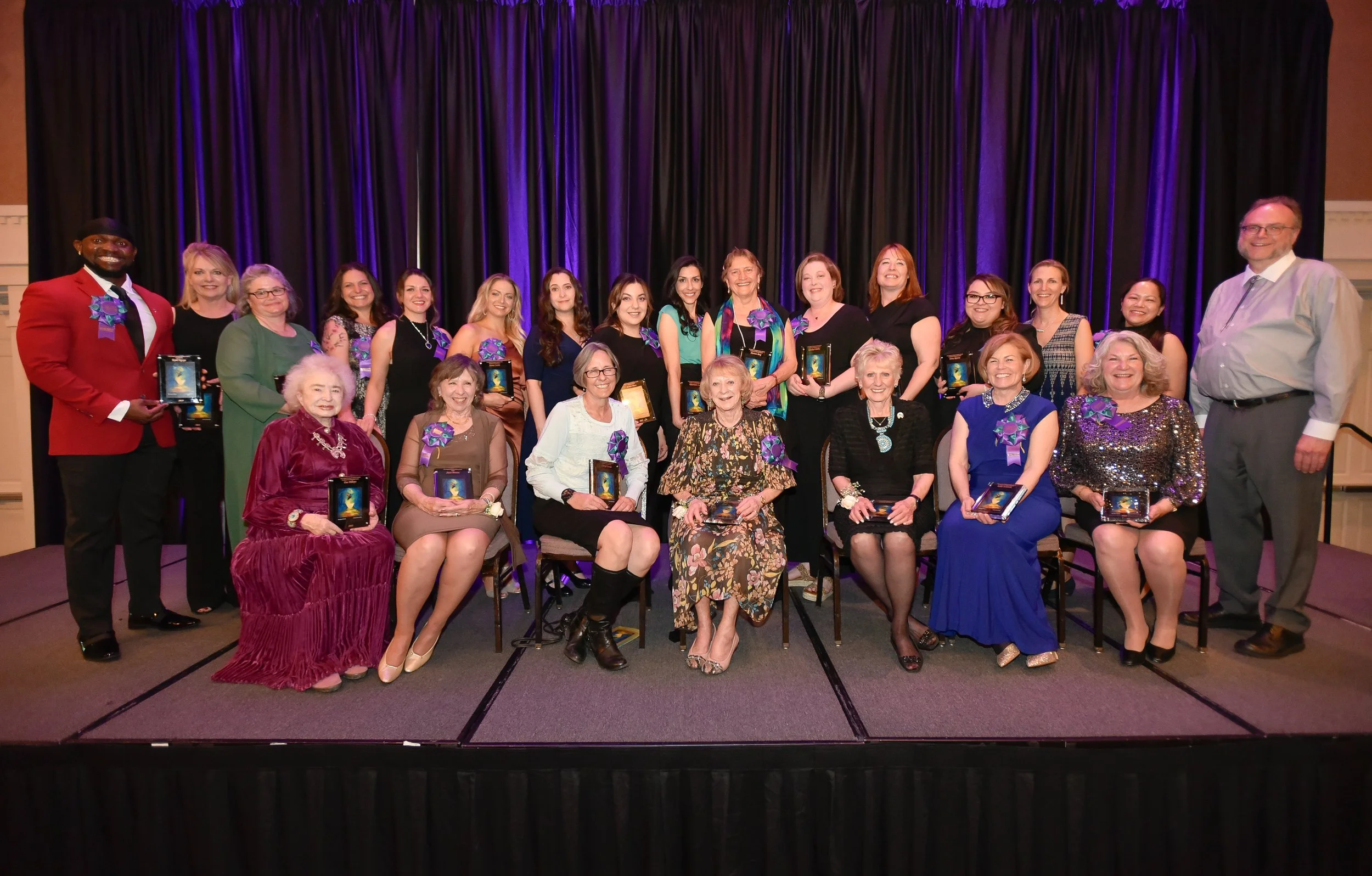 Group of people posing on a stage with awards, dressed formally, in front of a curtain with purple lighting.