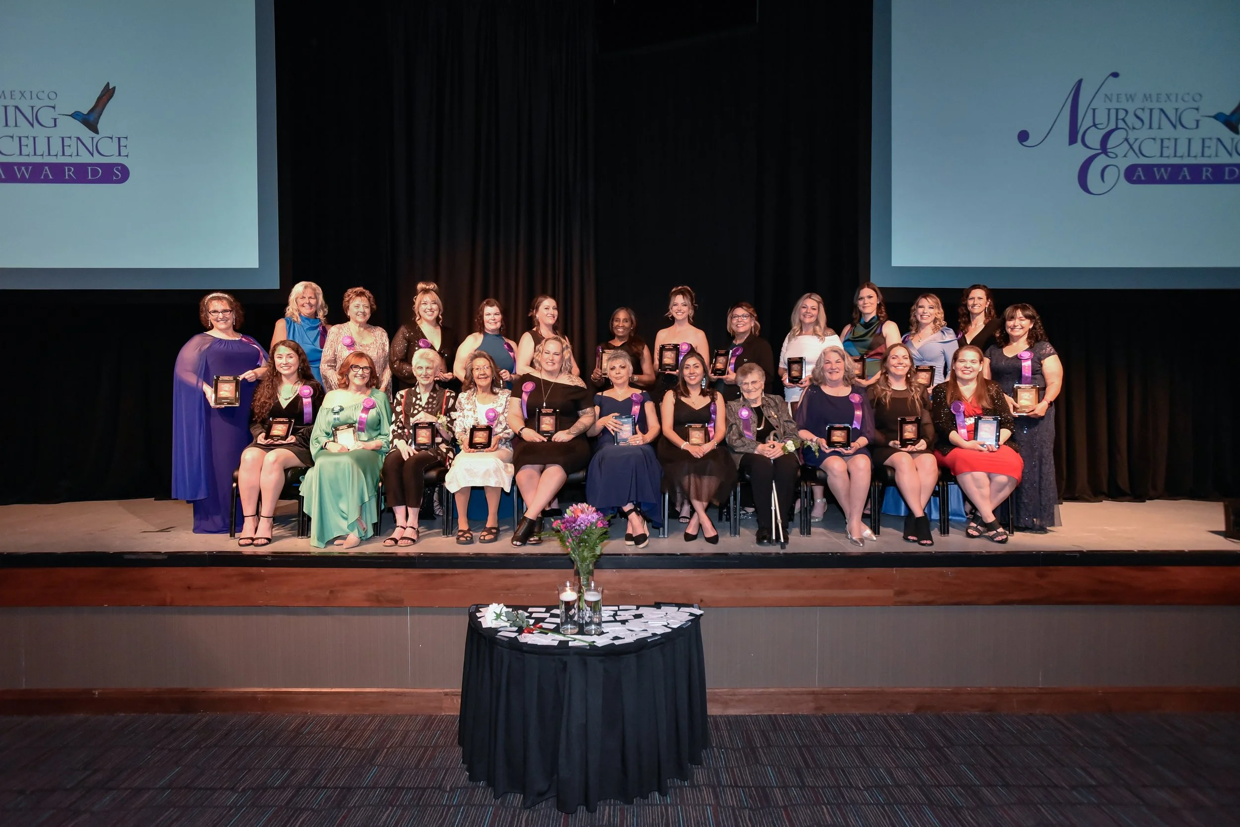 Group of women at an award ceremony on stage, holding awards, with a large screen displaying 'New Mexico Nursing & Excellence Awards' in the background.