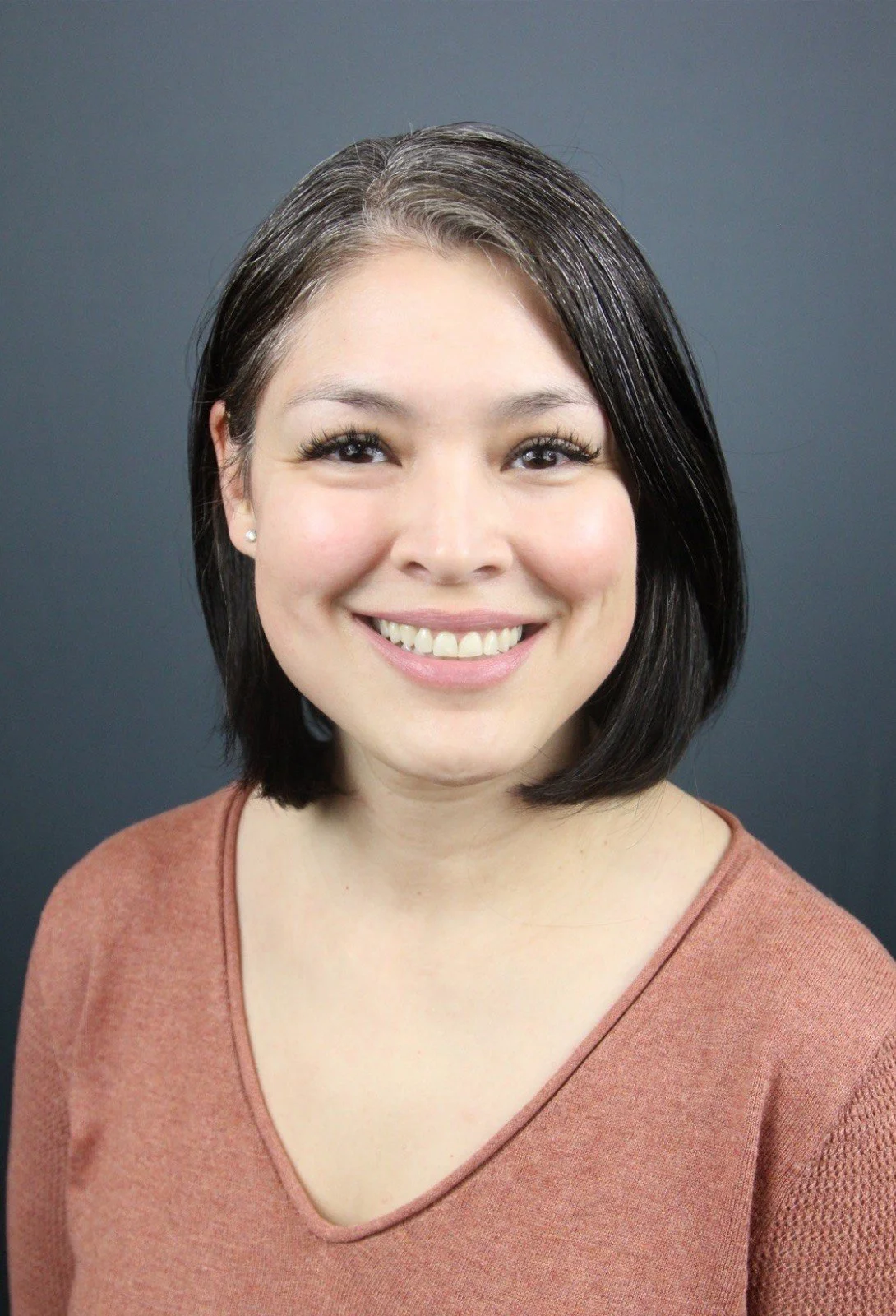 A woman with black hair and a warm smile, wearing a brown top, standing against a dark background.