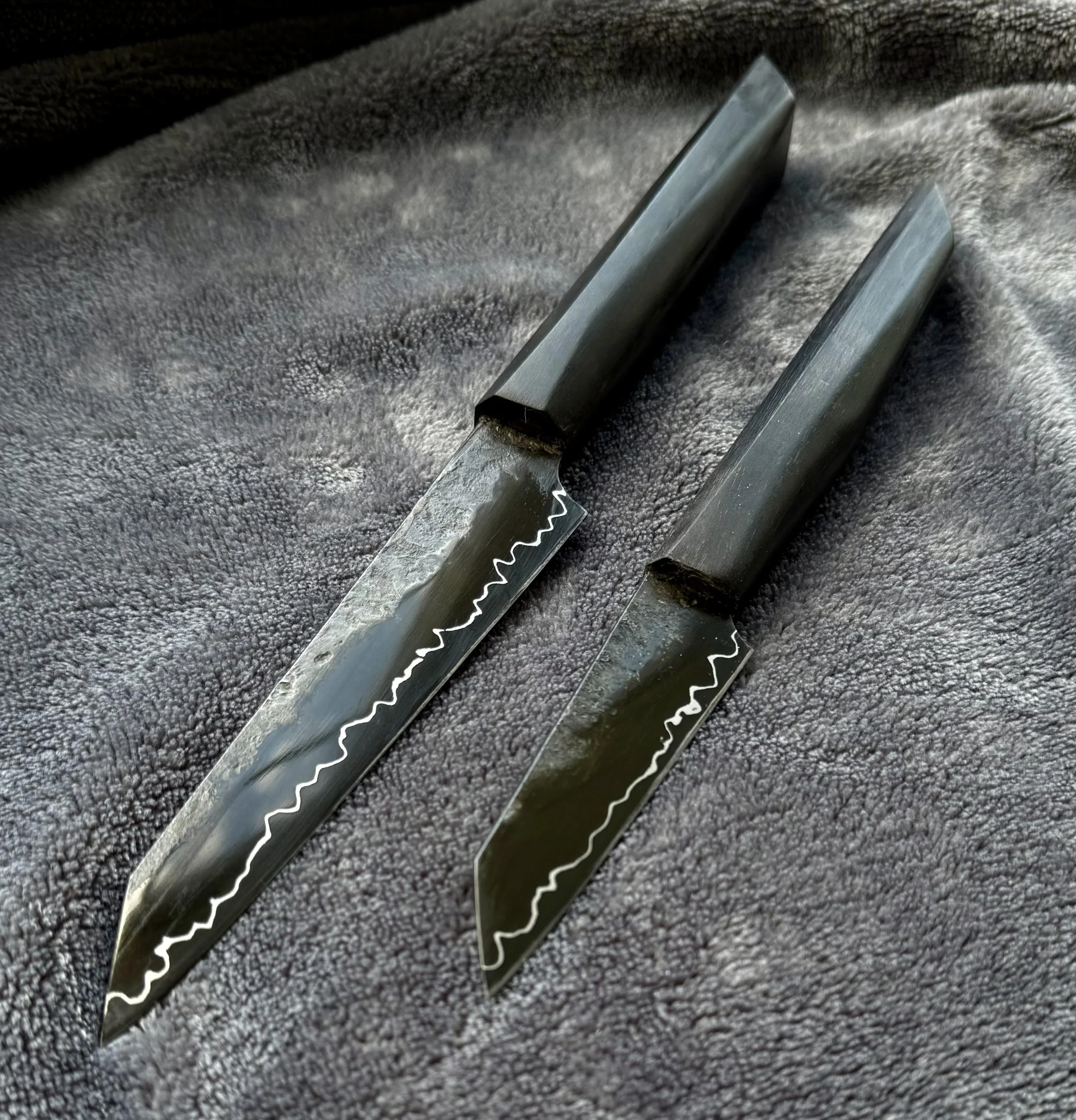 Two black-handled kitchen knives with jagged silver blades lying on a textured dark gray surface. pairing and petty knives