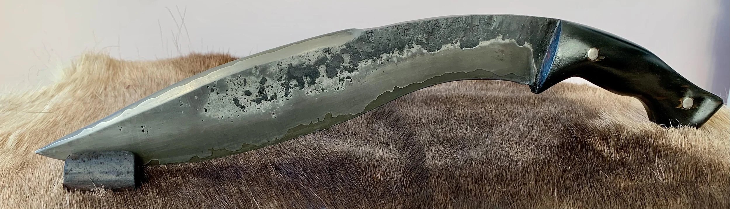 A large, vintage hunting or skinning knife with a black handle, placed on an animal hide with brown fur. Kukri