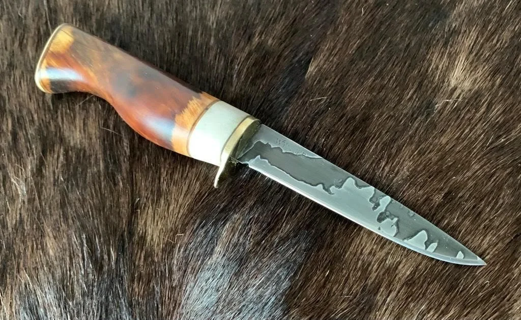 A handcrafted hunting or kitchen knife with a curved handle made from multicolored wood and a Damascus steel blade, resting on a dark fur background.