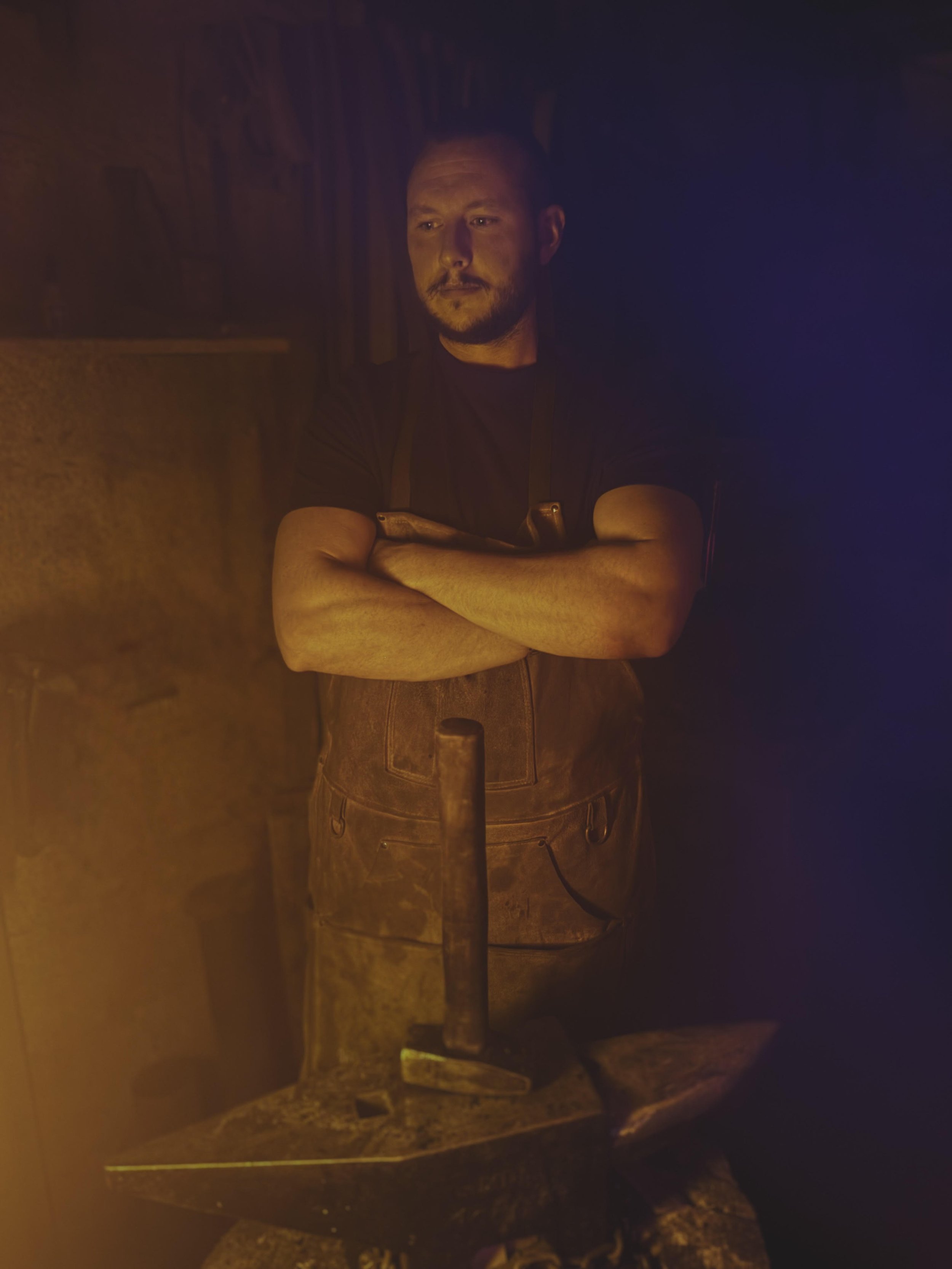 A blacksmith standing with arms crossed in a dimly lit workshop, with a hammer and anvil in front of him.