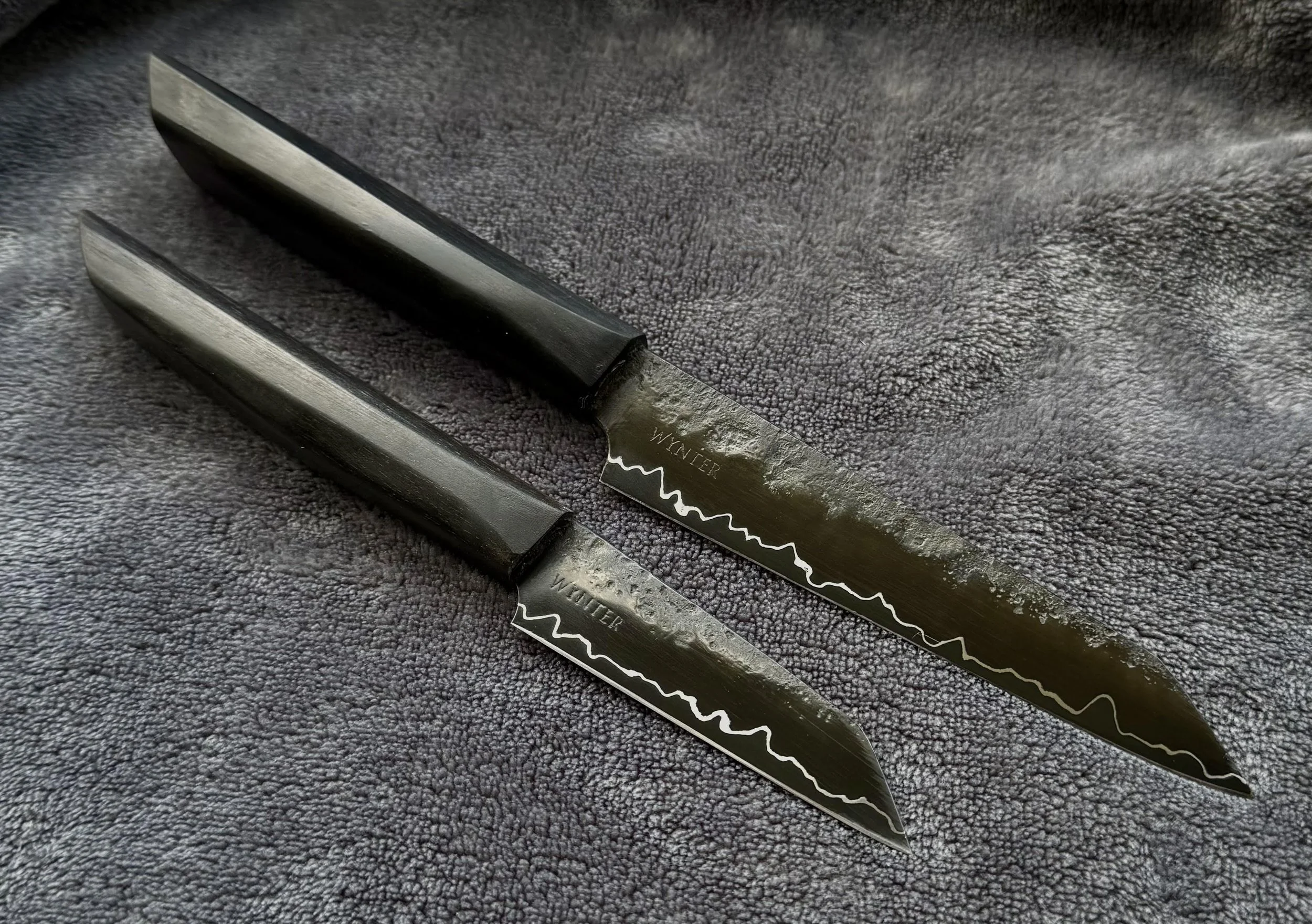 Two black-handled kitchen knives with black blades lying on a textured, dark gray surface.