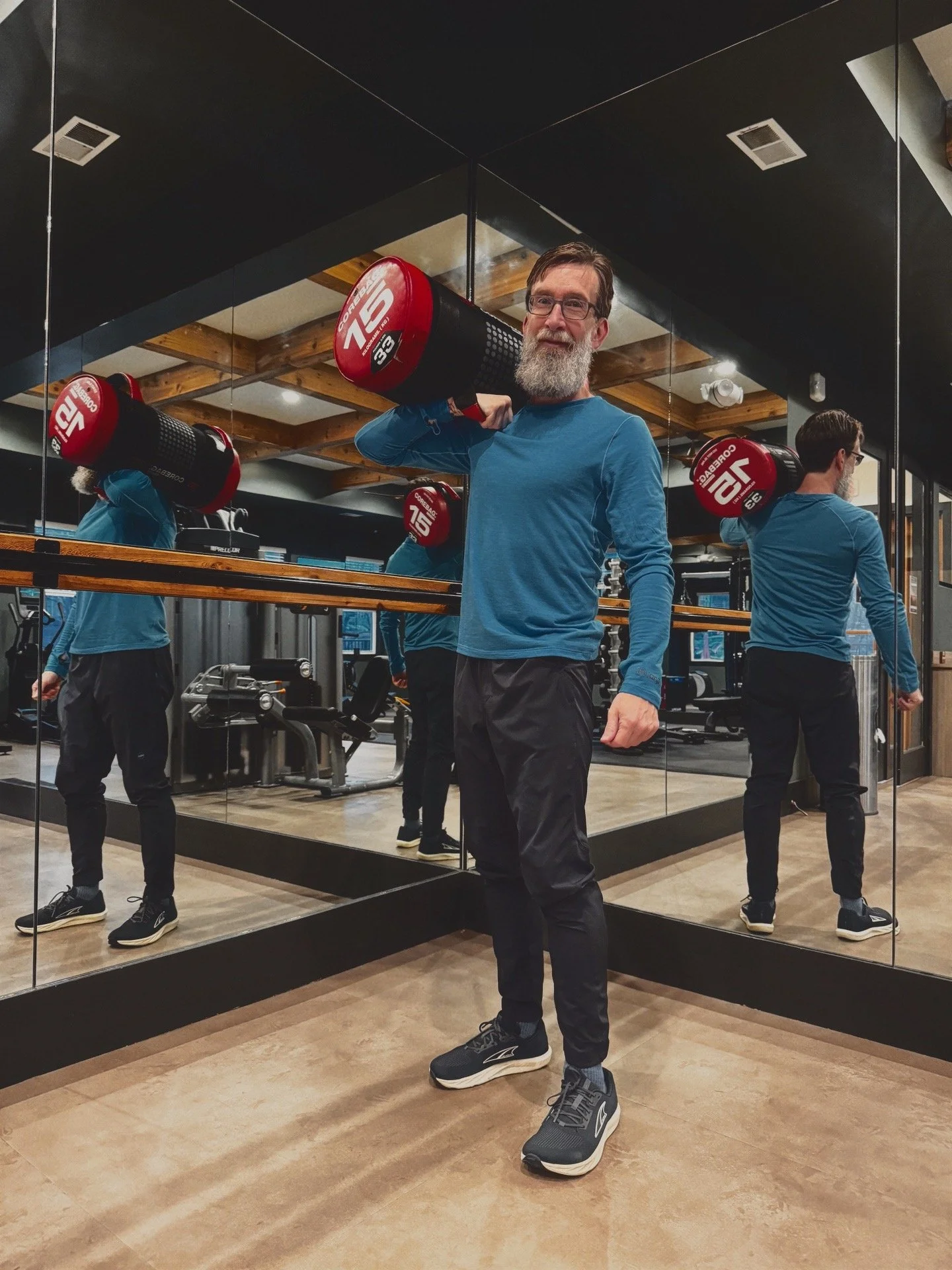 Tim Earley in an indoor gym smiling at the camera while carrying a 33 pound sandbag on his shoulder.
