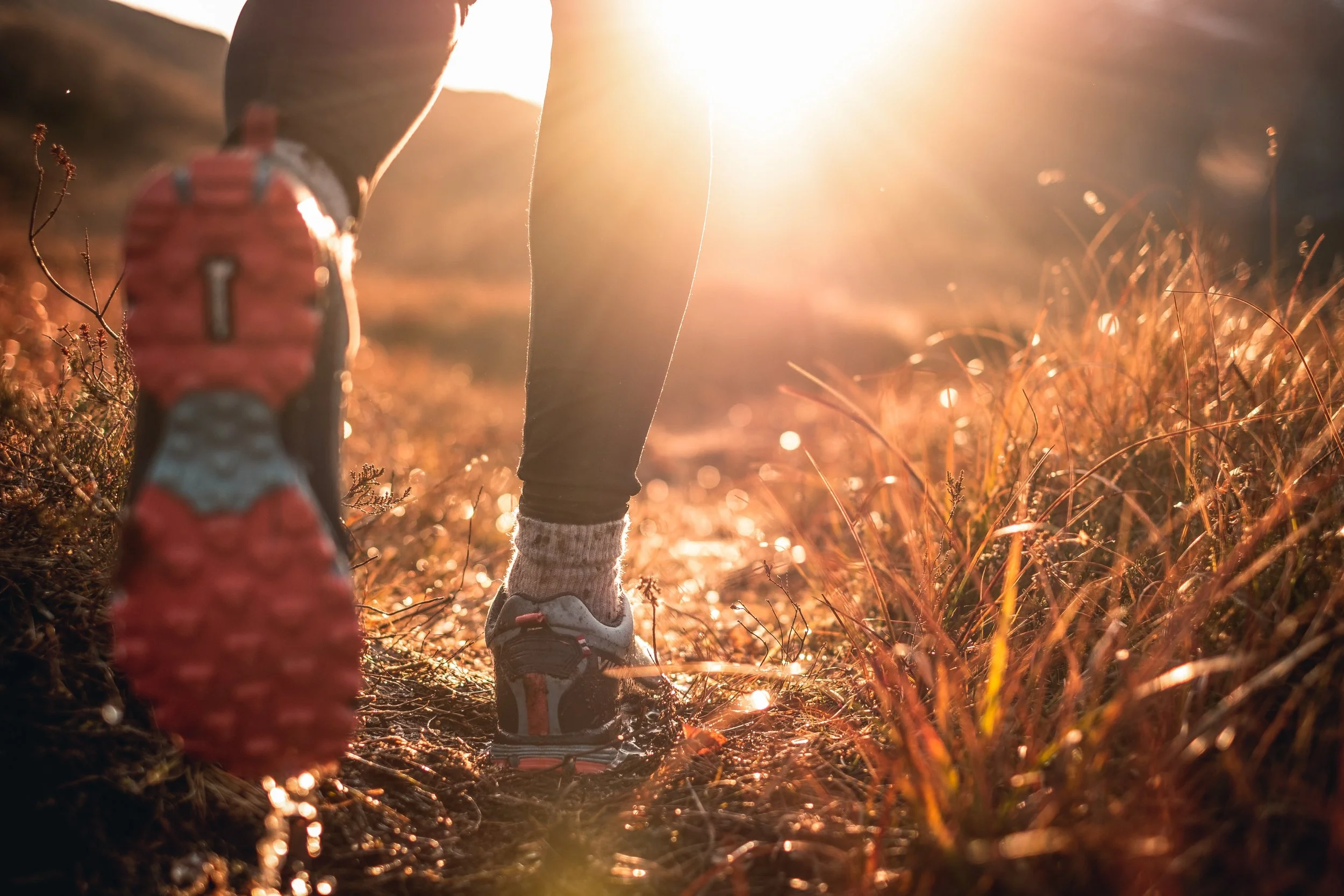 Close-up of trail running shoes on a dirt path at sunrise, runner stepping through grass with sunlight flaring ahead of them, symbolizing outdoor fitness, hiking, and a healthy lifestyle.