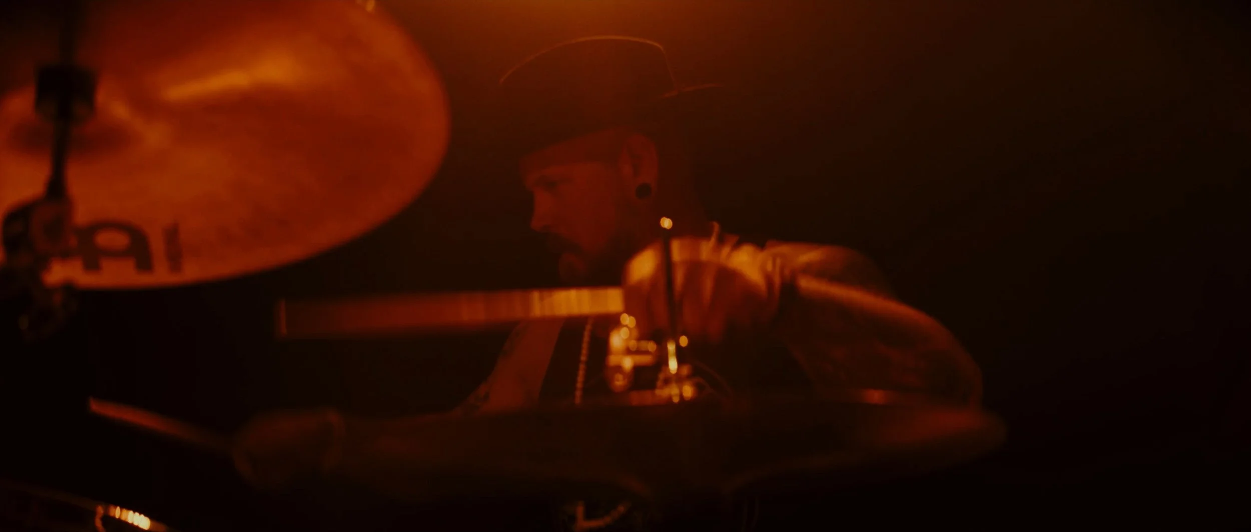 A male drummer with tattoos, wearing a backward cap, playing drums in a dimly lit, orange-hued environment.
