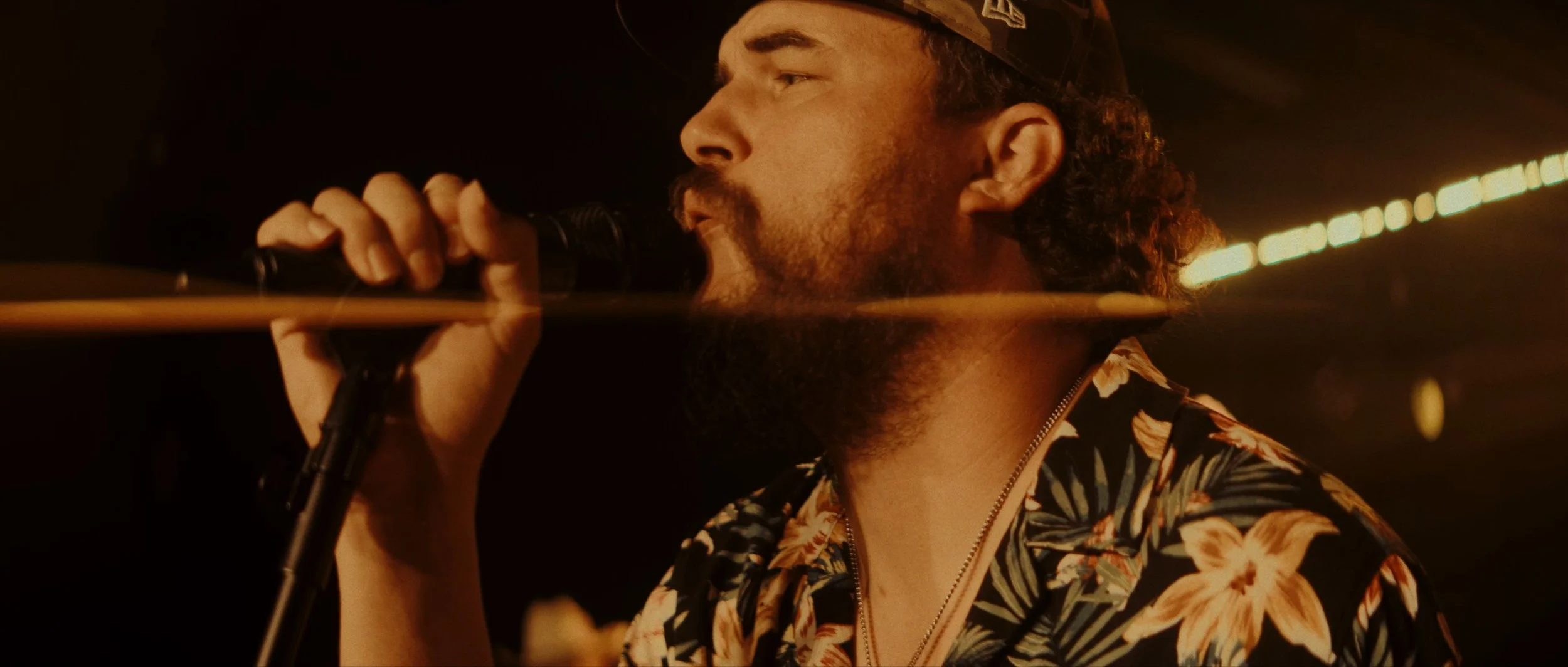 Man singing into a microphone, wearing a floral shirt and a cap, with warm lighting and a dark background.