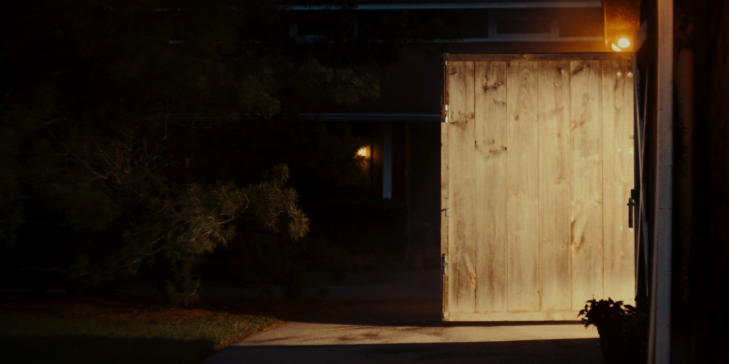 Nighttime view of an open garage door with a lit interior, wooden panels, and trees outside in the dark.