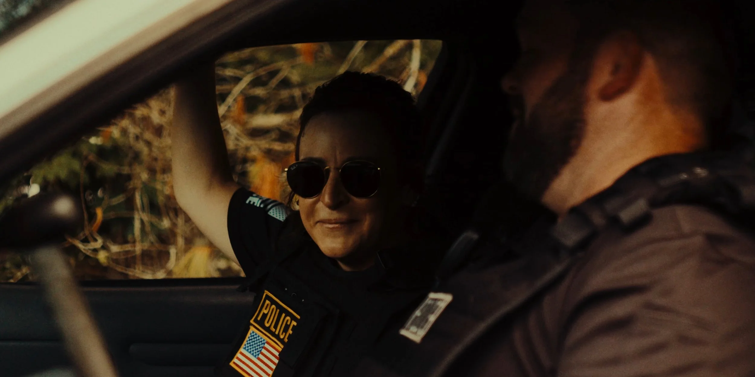 A police officer talking to a man inside a vehicle during daytime. The officer is wearing sunglasses and a black uniform with a police patch and American flag badge, and appears to be smiling.