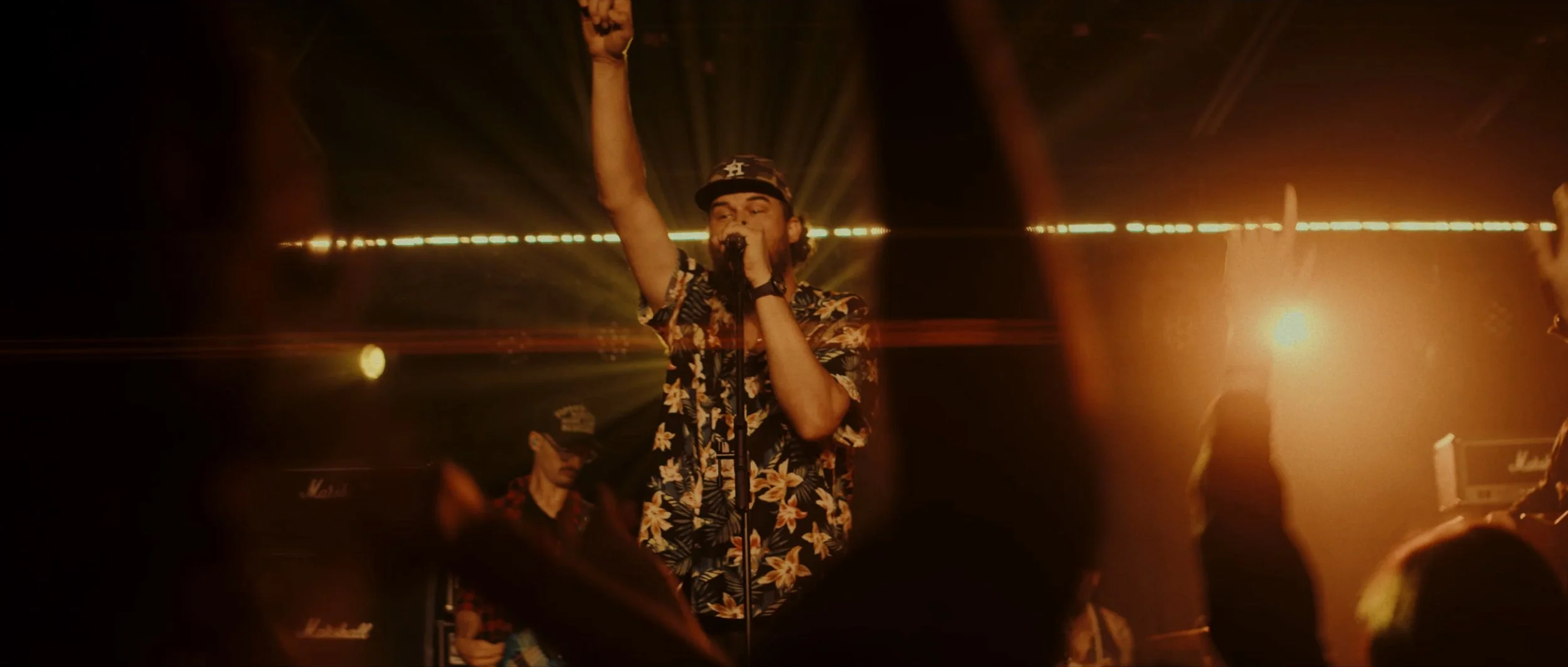A man wearing a floral shirt and baseball cap singing into a microphone on stage, backlit with warm yellow lights, with an audience in front