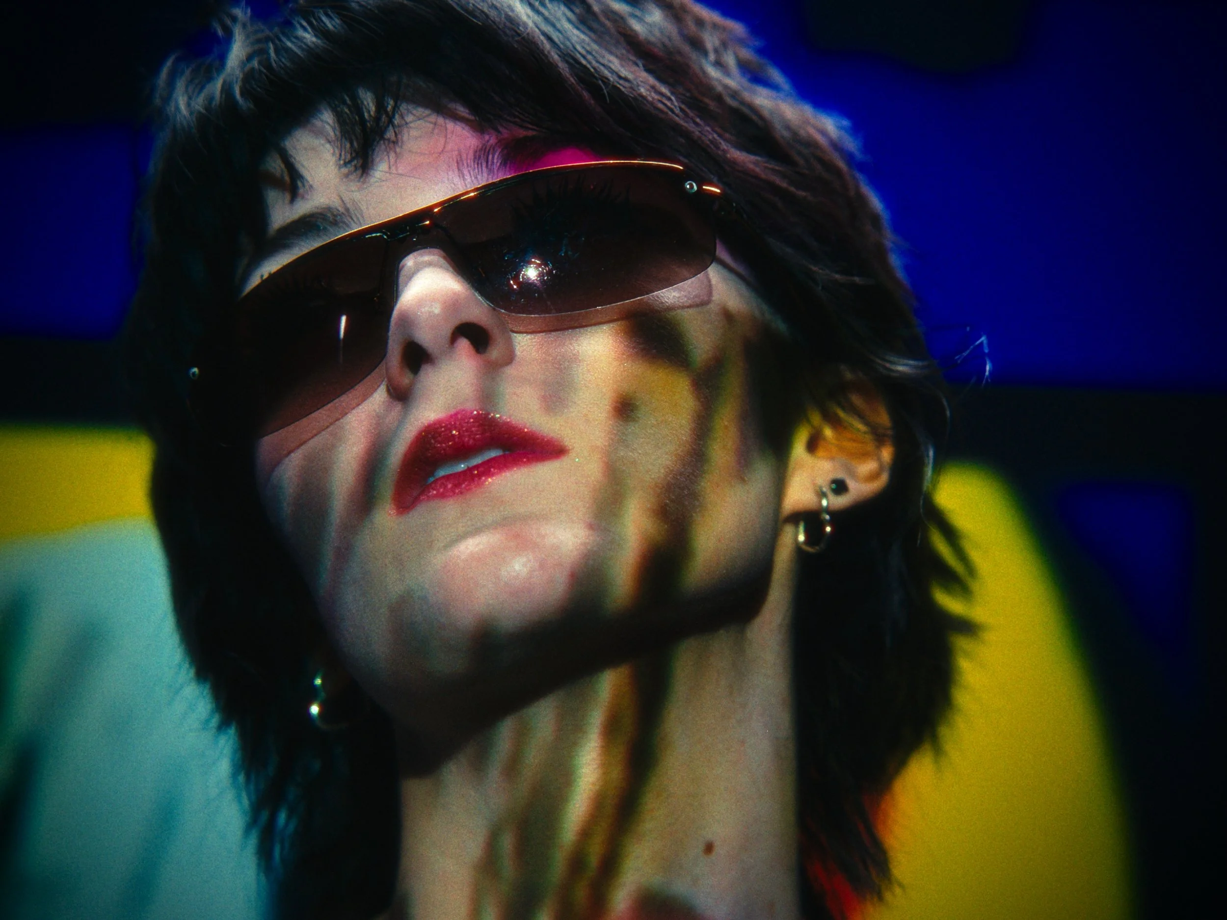 A woman wearing sunglasses and earrings, with short dark hair and red lipstick, is captured in a close-up photograph. The image has colorful lighting effects.