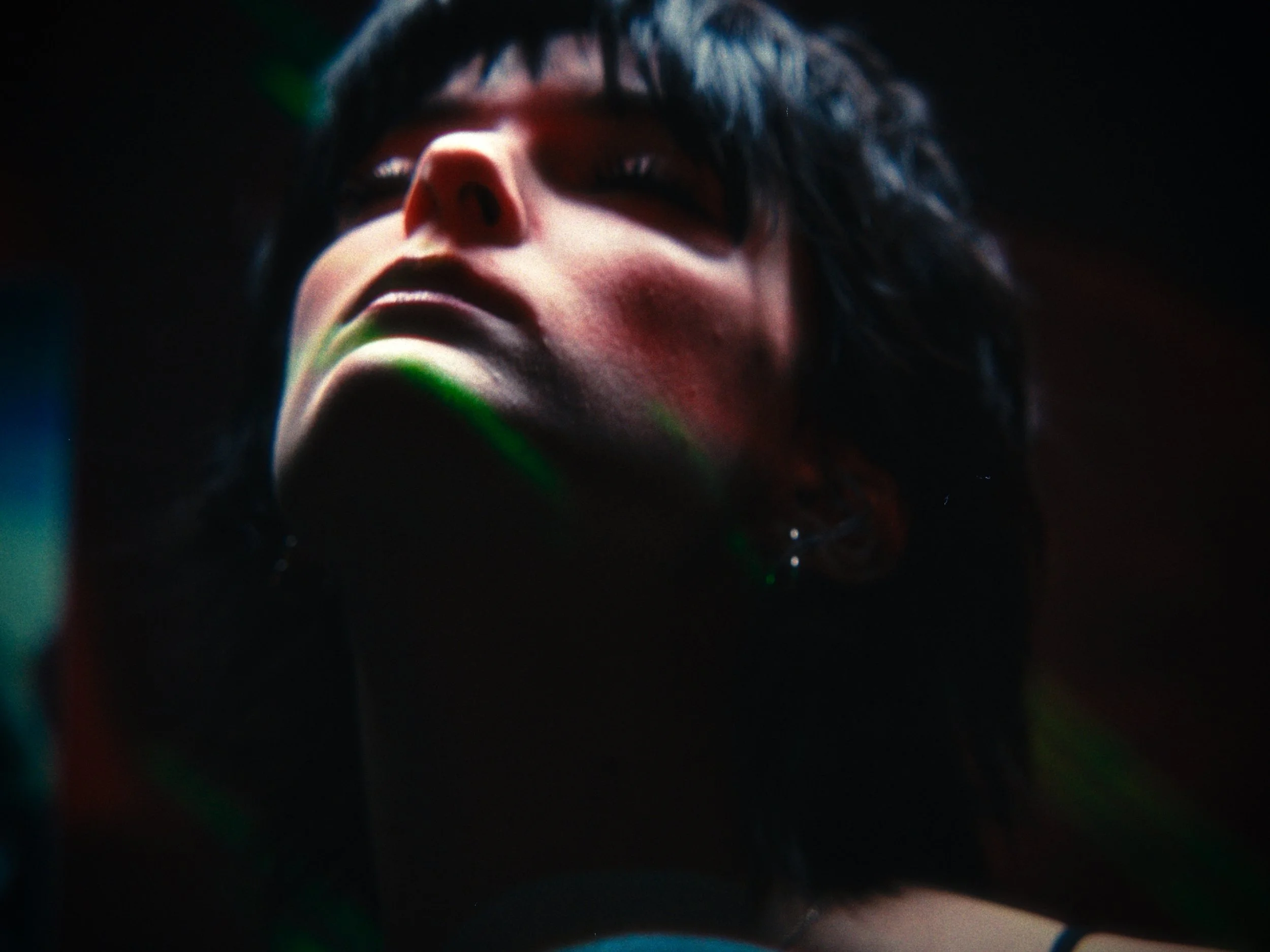 A close-up of a person with dark, short hair, wearing sunglasses, with their head tilted slightly back in a dark environment with colorful light streaks.