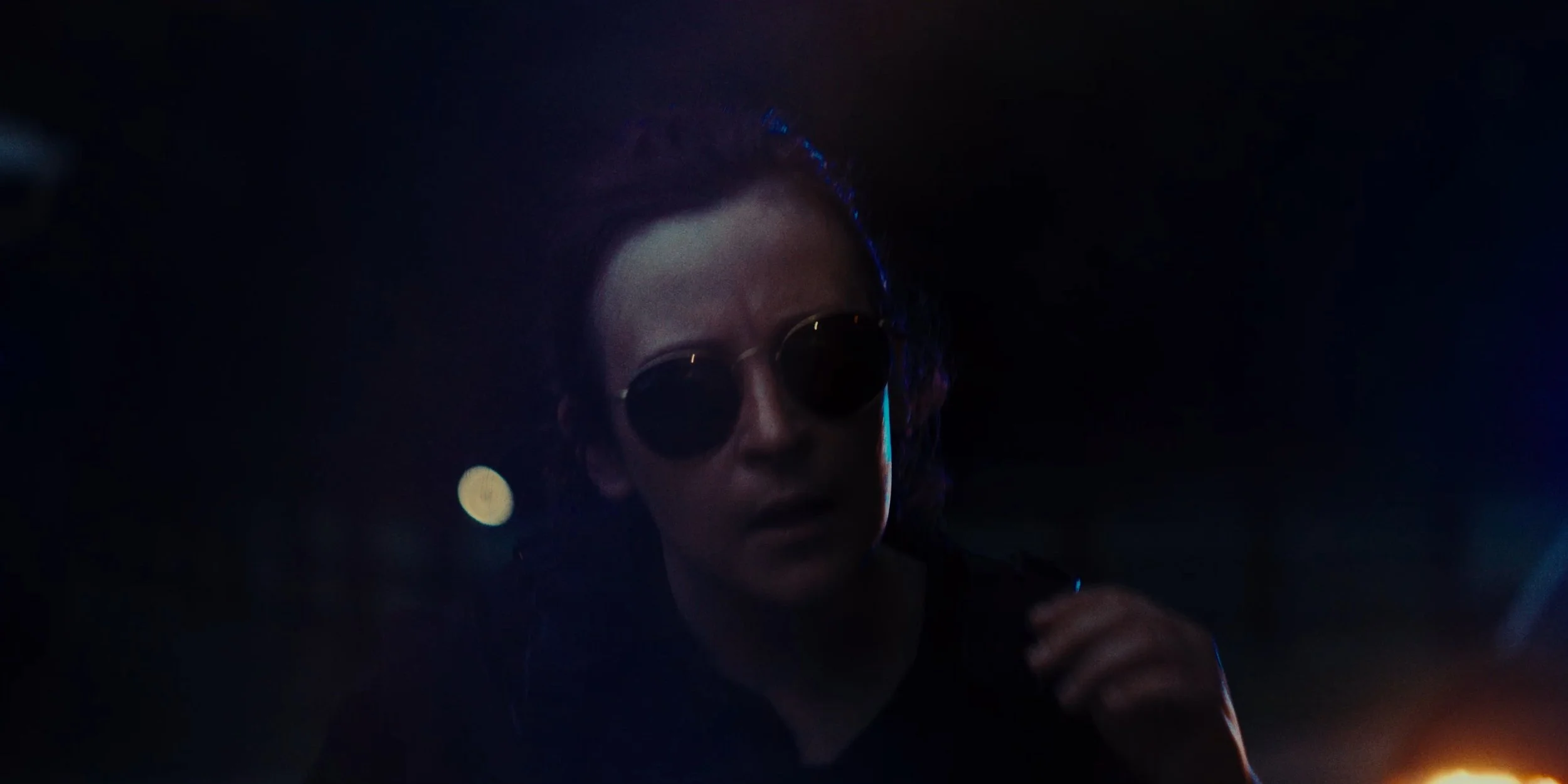 Person wearing dark sunglasses in a dark environment with subtle lighting and reflections.