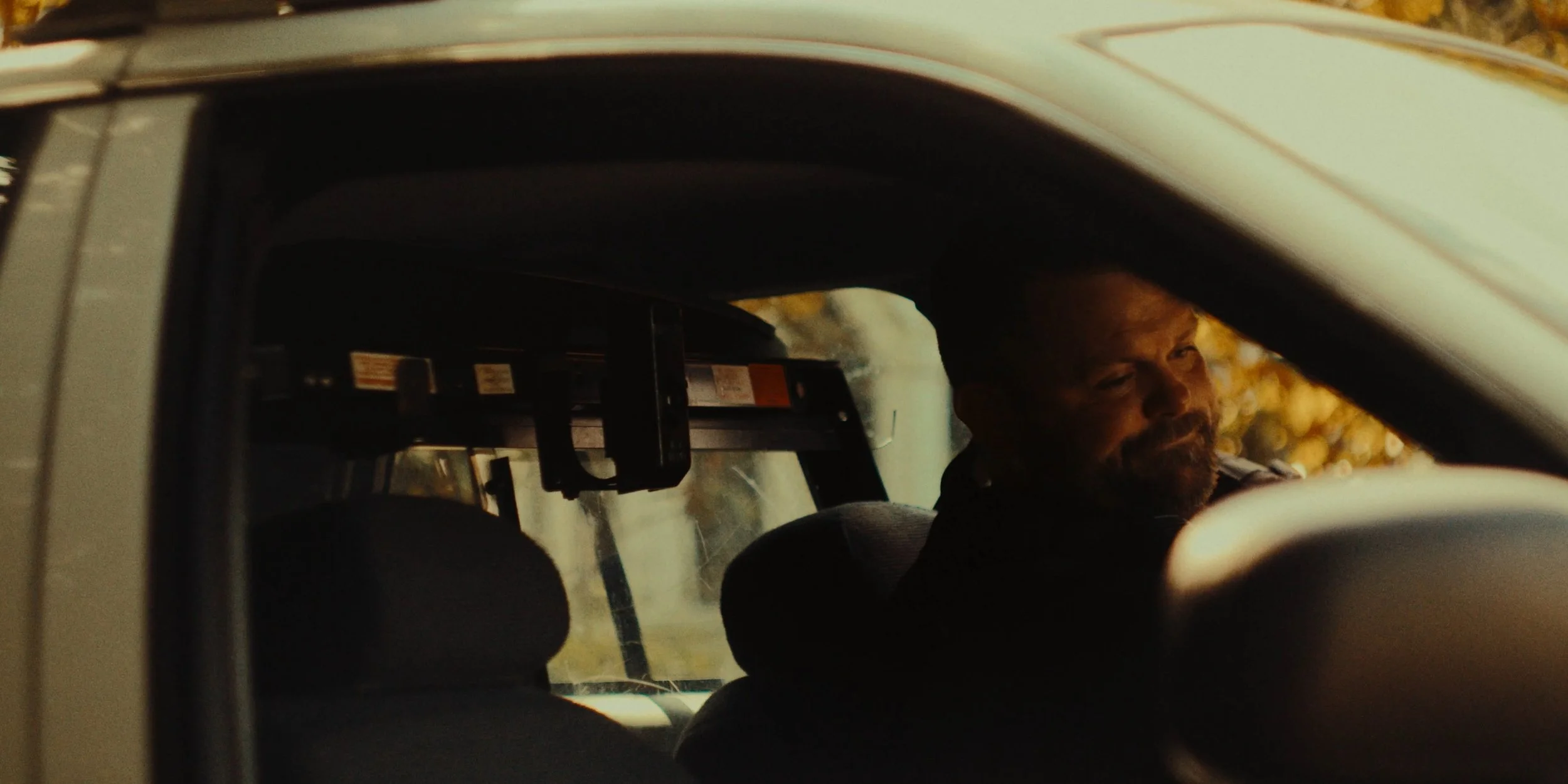 A man with a beard driving a vehicle, seen through the driver's side window in warm lighting.