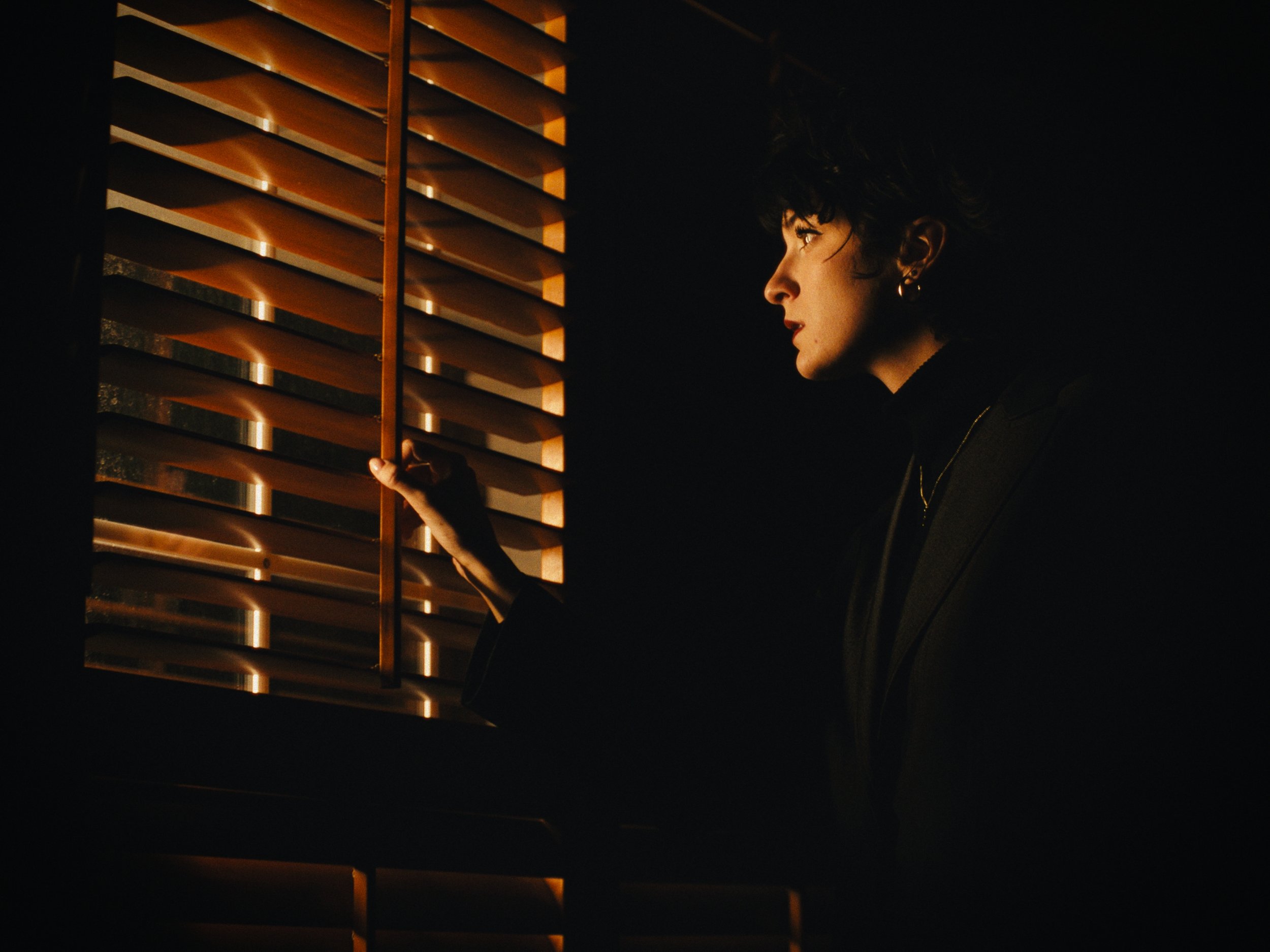 A woman with short curly hair and earrings gazes through window blinds at night, illuminated by warm light.