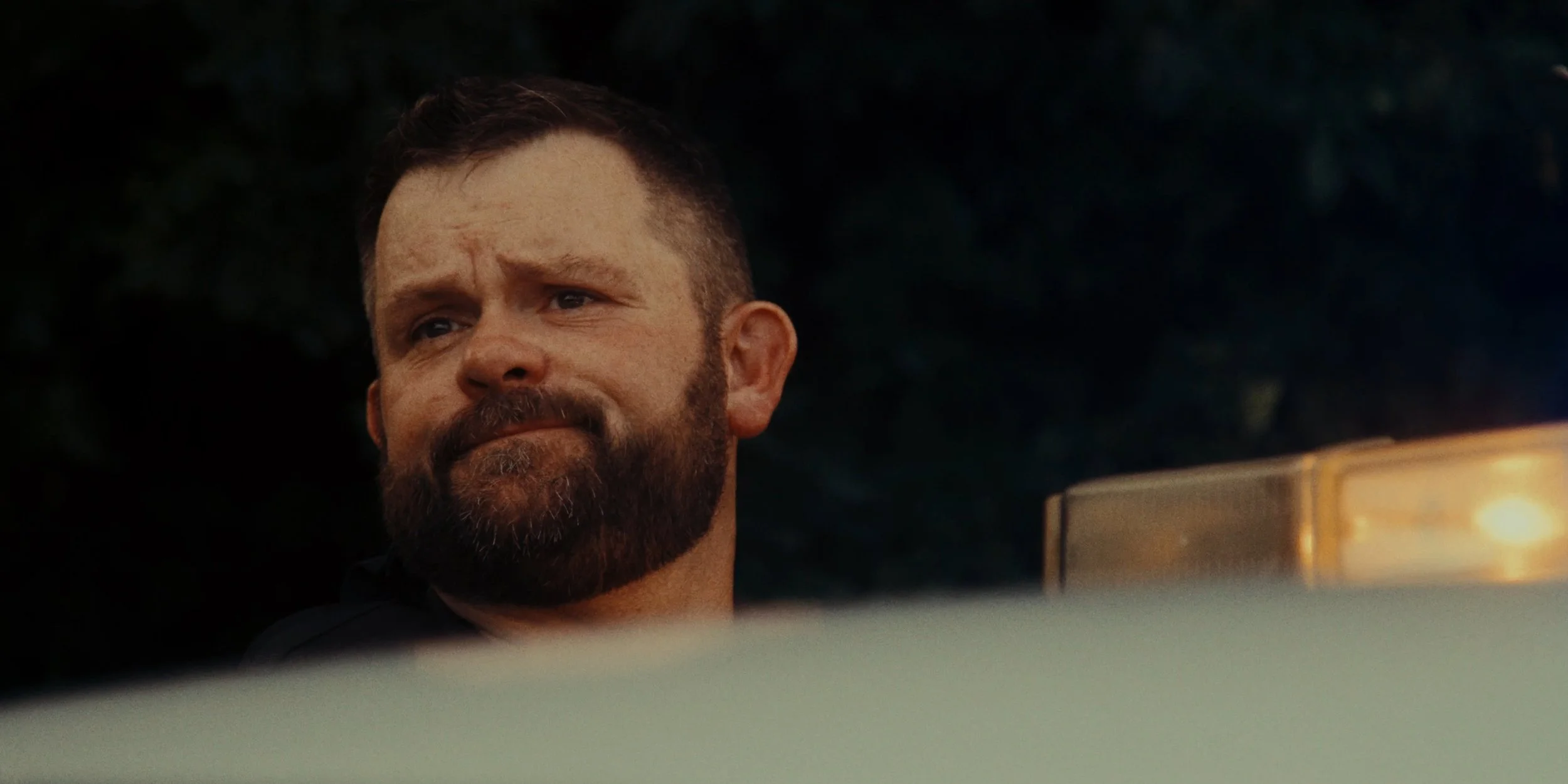 A man with a beard and short hair in a dark background, looking over a white surface, possibly a vehicle.