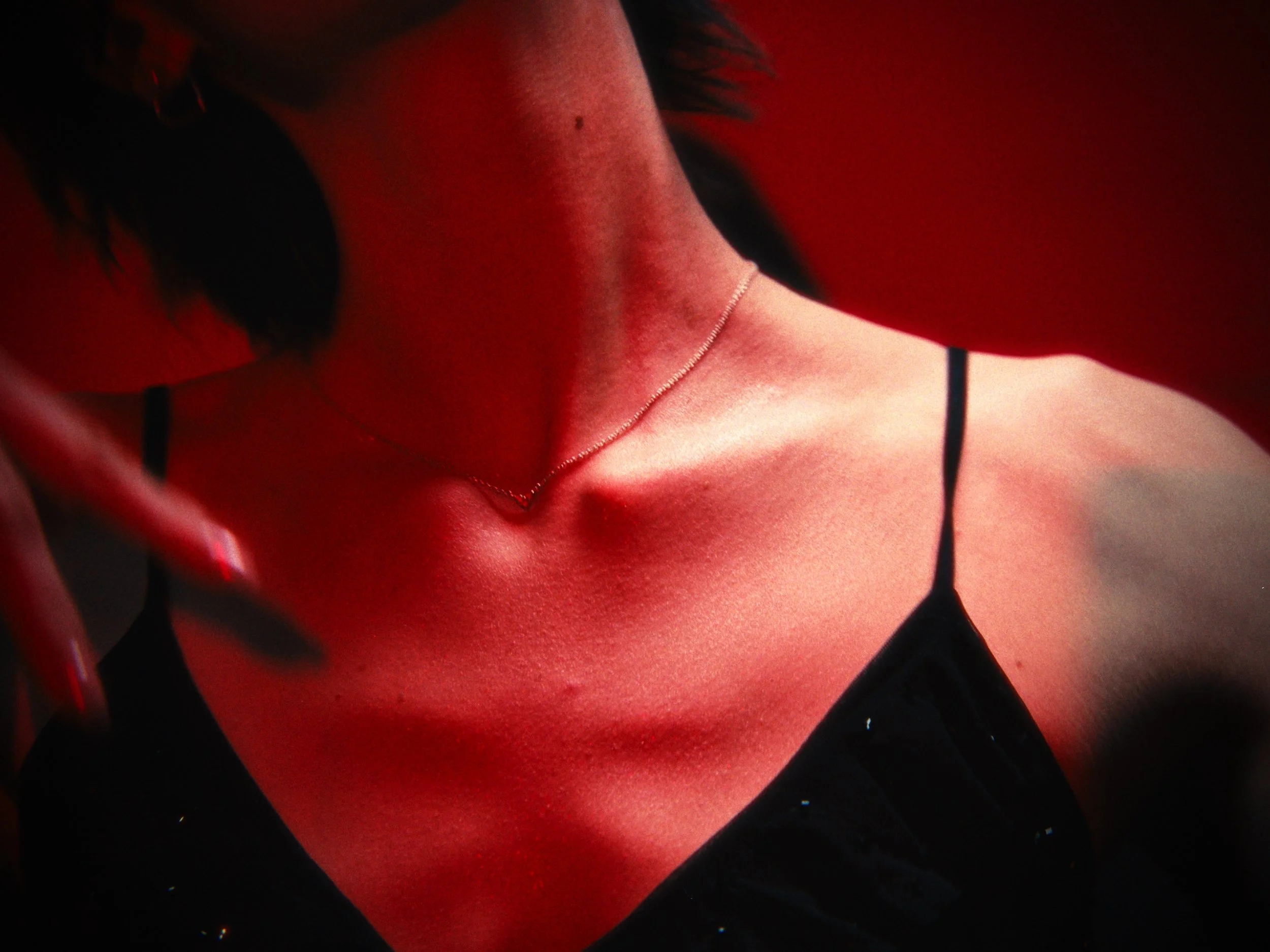 Close-up of a woman's neck and chest area illuminated with red lighting, wearing a thin necklace and a black spaghetti strap top.