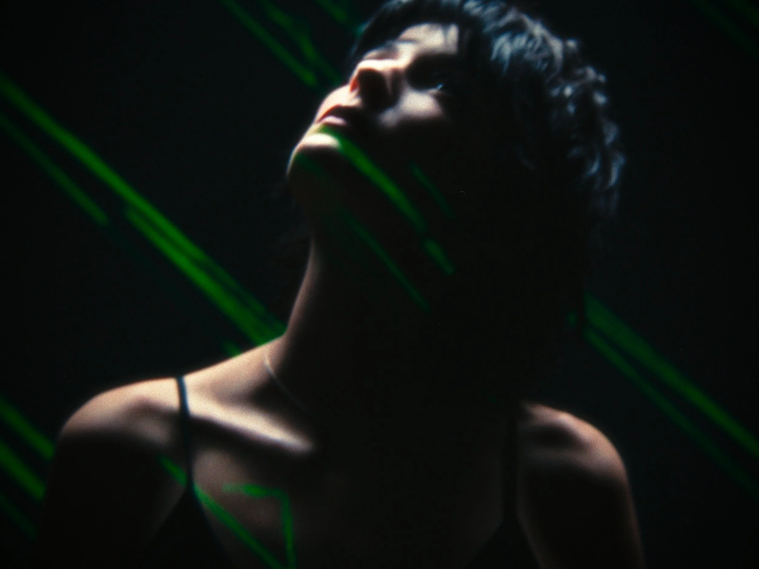 A woman with curly hair looking upward in a dark setting with green laser beams crossing in front of her.