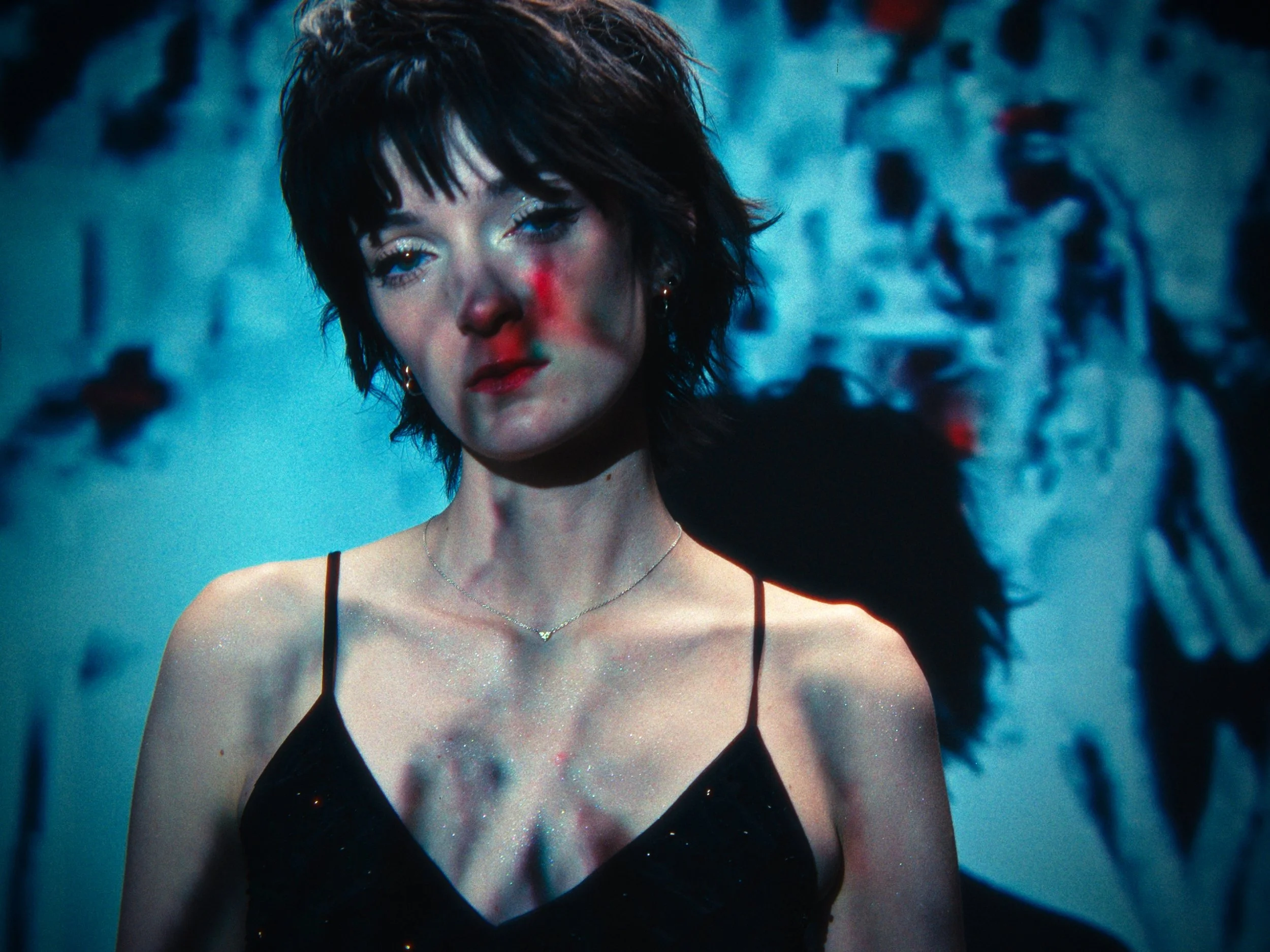 A young woman with short dark hair and a tear-streaked face, wearing a black spaghetti strap top, standing against a blurred background with faint figures.