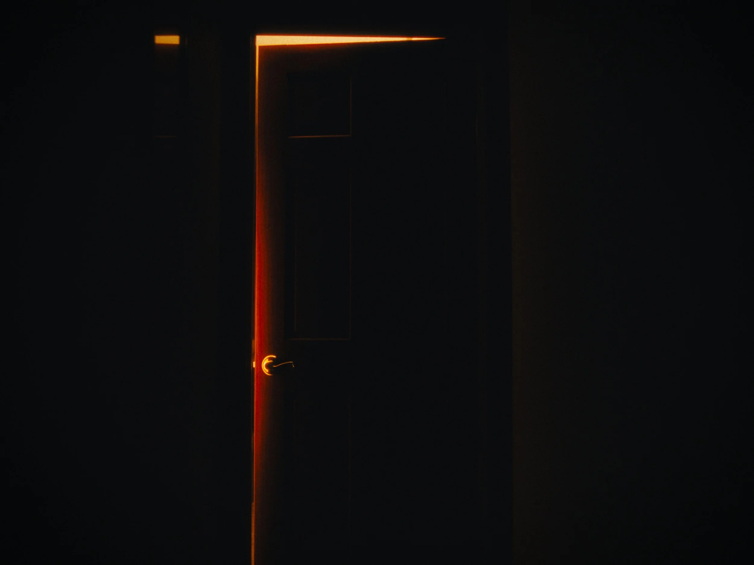 A dark room with a partially opened black door, illuminated by a warm orange light along the door's edge and handle.