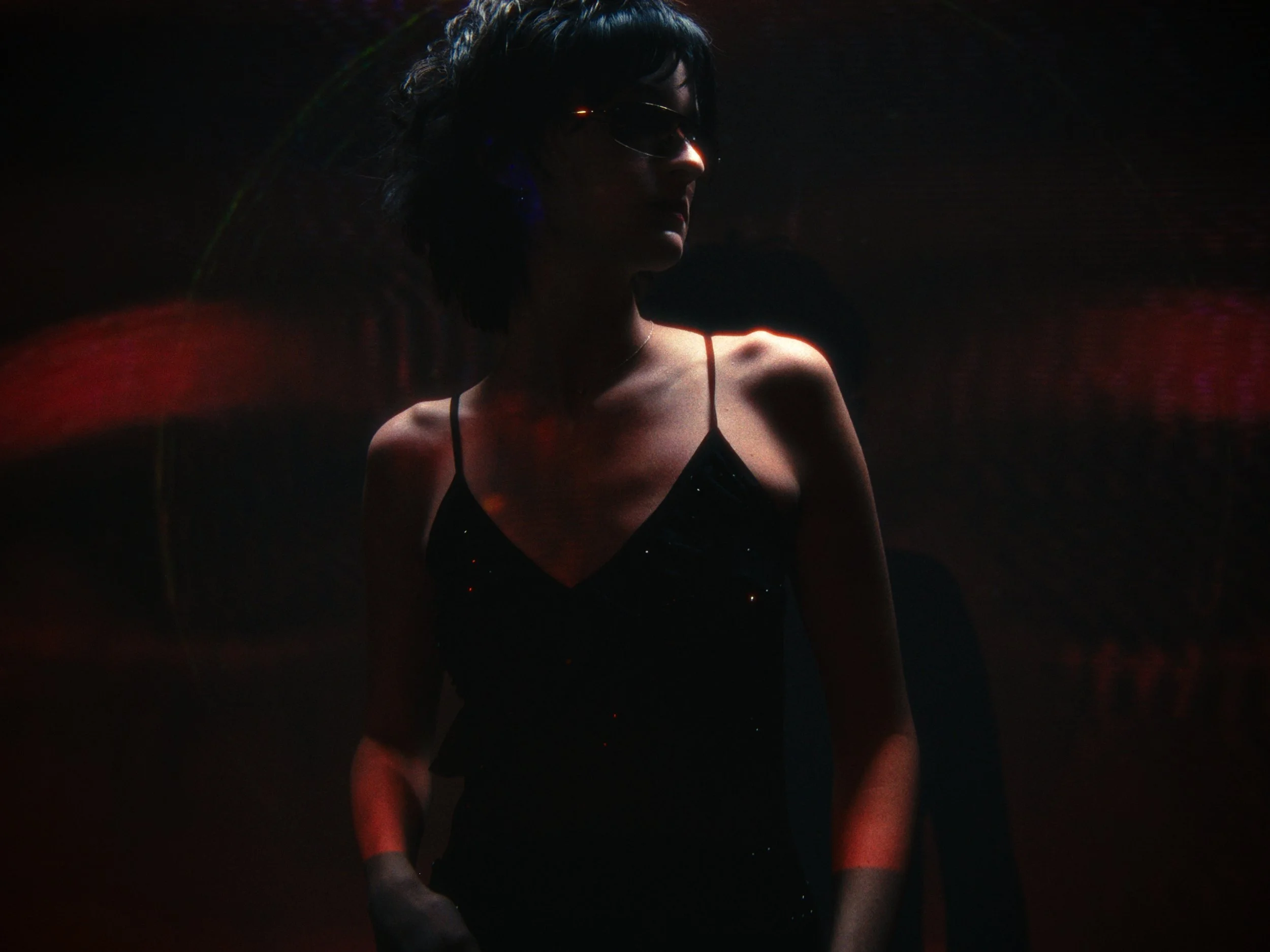 A woman with short dark hair wearing sunglasses and a black spaghetti strap dress, standing in a dark environment with low lighting and some red and purple light streaks.