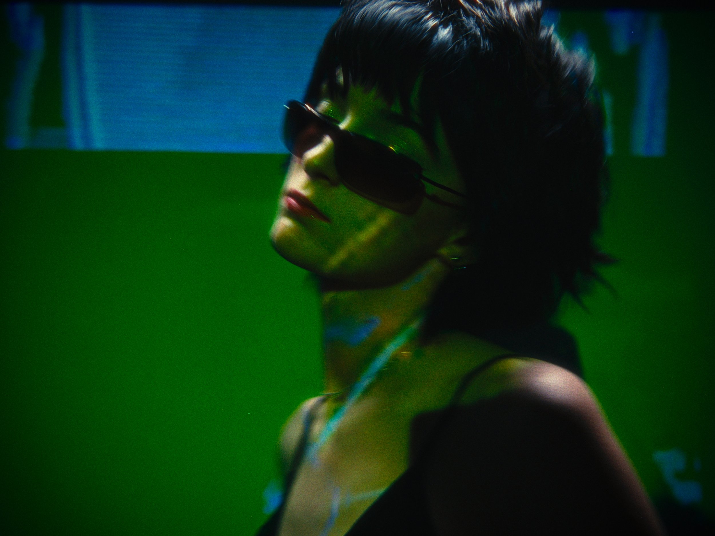 A woman wearing sunglasses with projection lights on her face and neck, against a green and blue background.