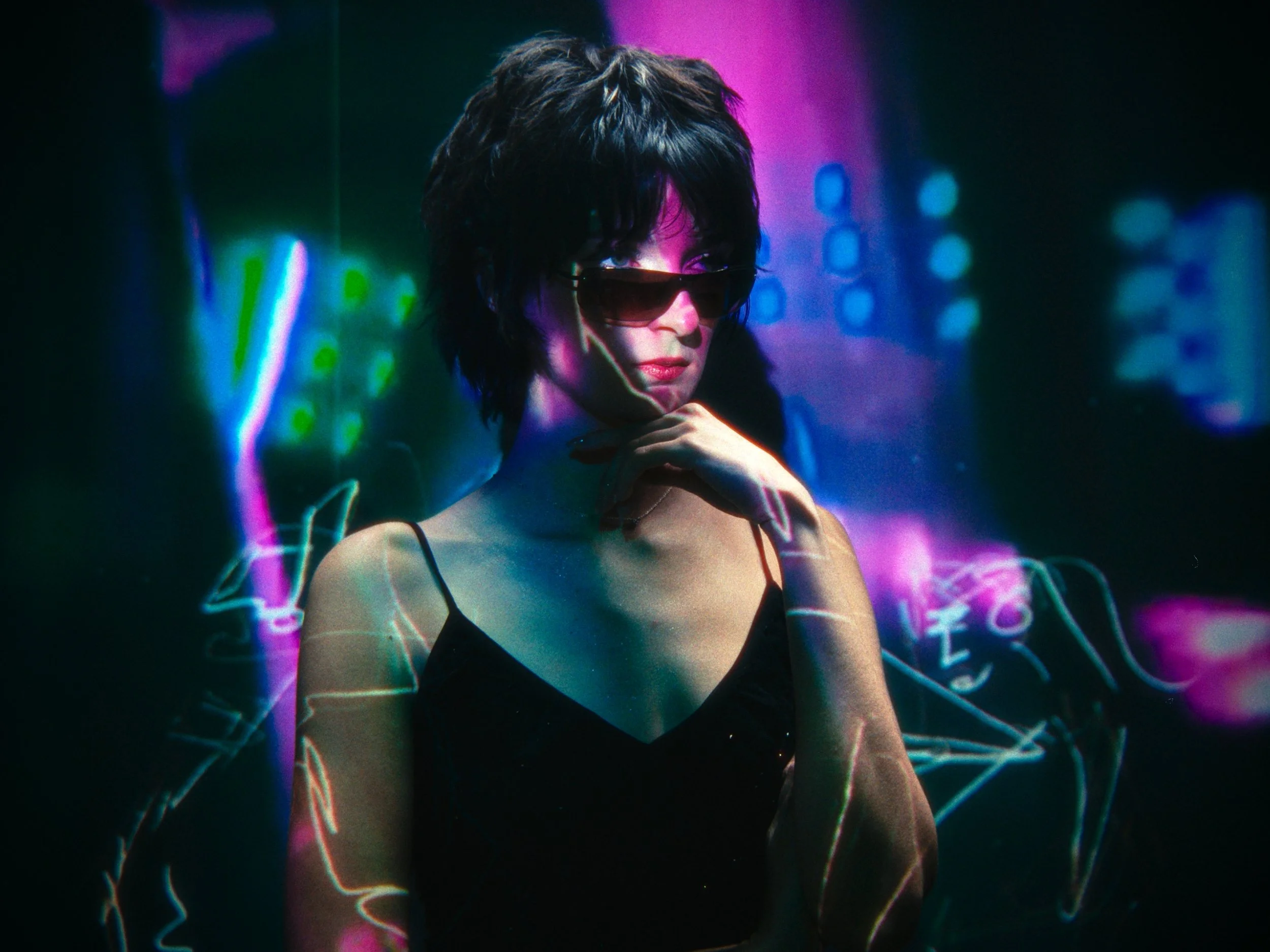 A woman with short dark hair wearing sunglasses and a black spaghetti strap top, illuminated by colorful neon lights and electronic music waveforms in the background.