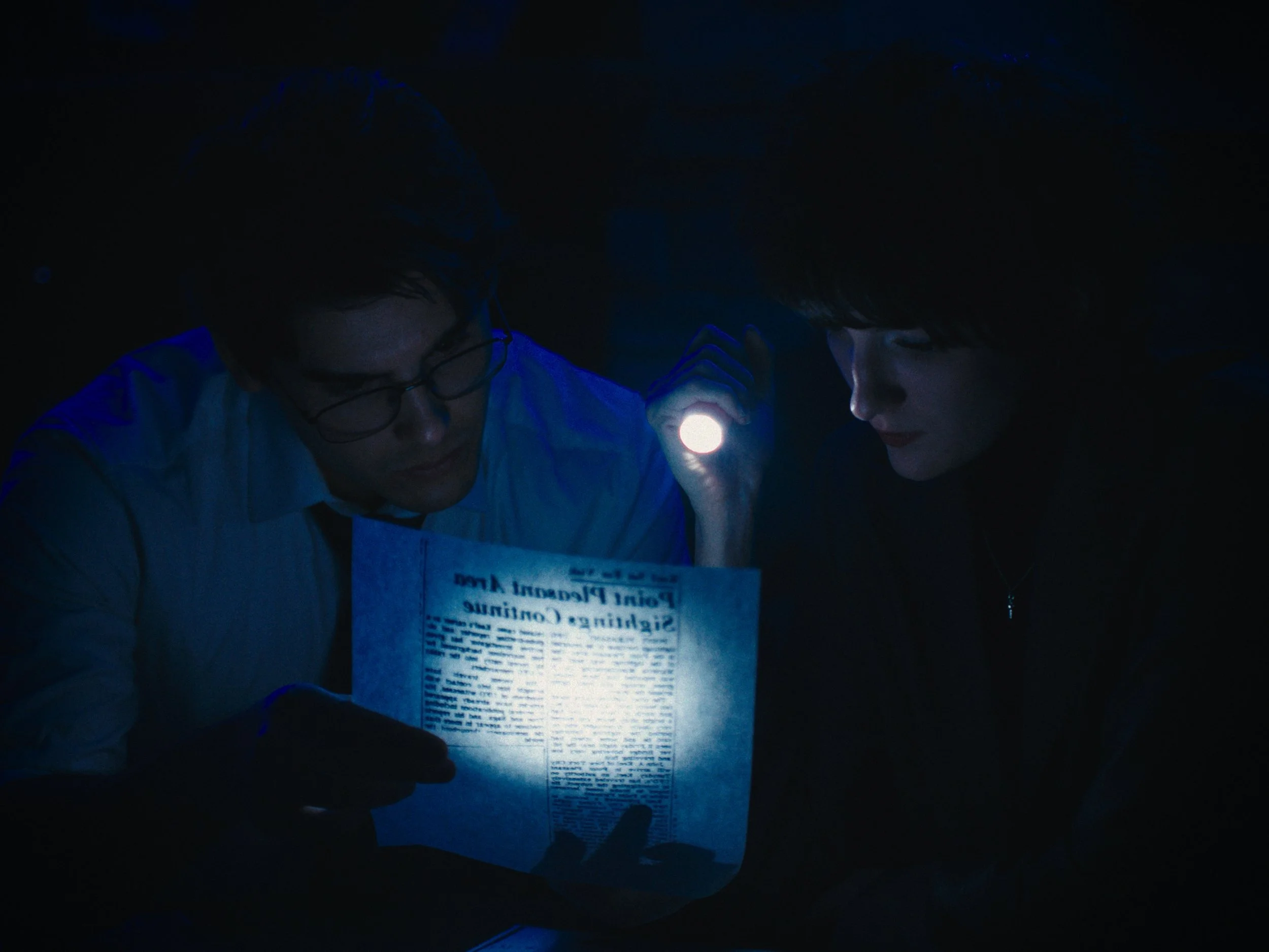 Two people illuminated by a flashlight in a dark room, examining a newspaper.