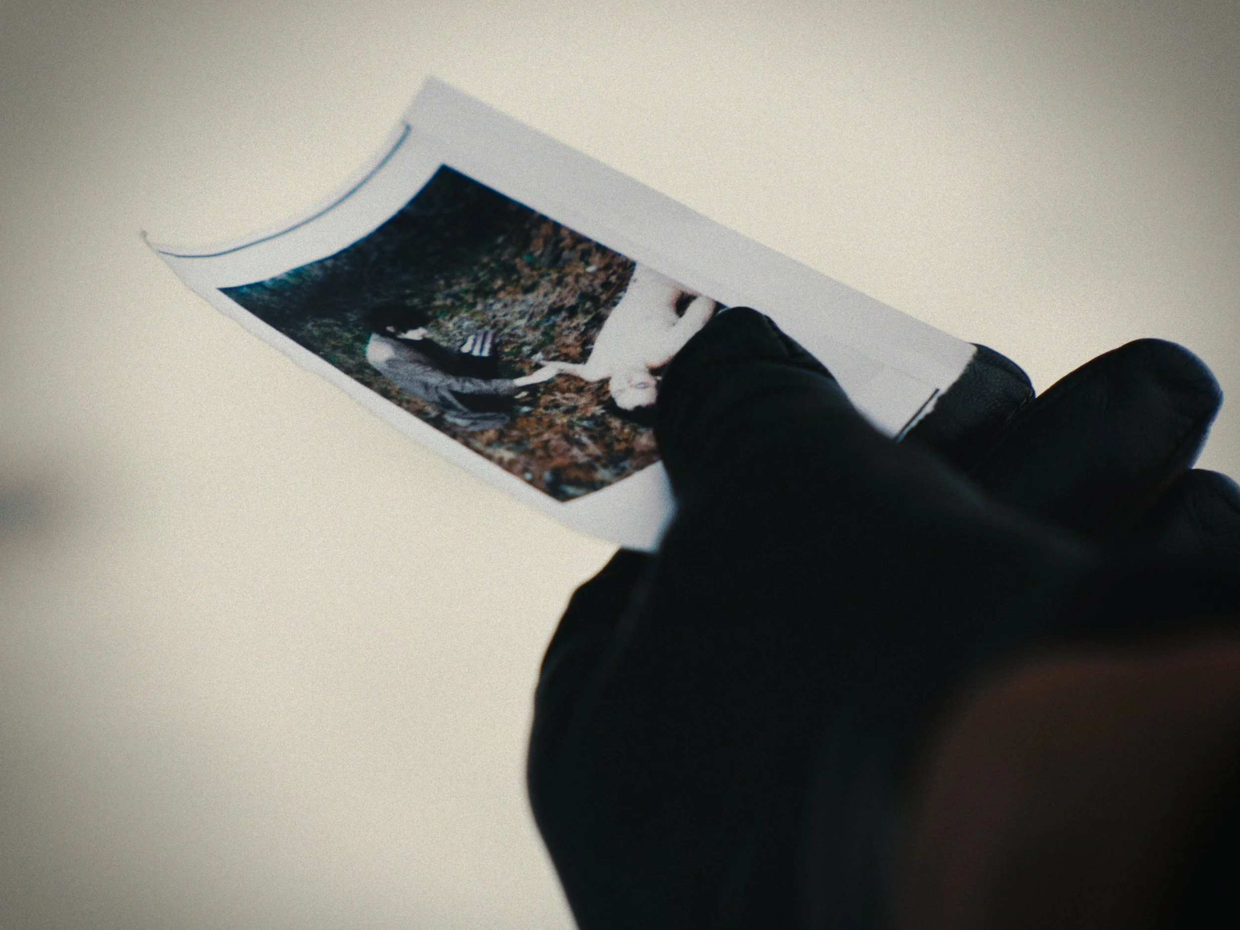 Person wearing black gloves holding a printed photograph of someone lying on the ground outdoors.