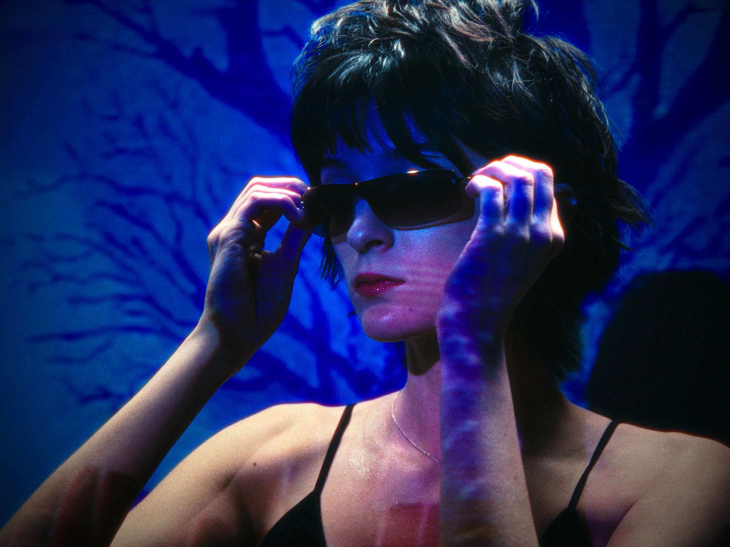 A woman with short dark hair wearing sunglasses and a black spaghetti strap top, adjusting her sunglasses under a blue lighting background.