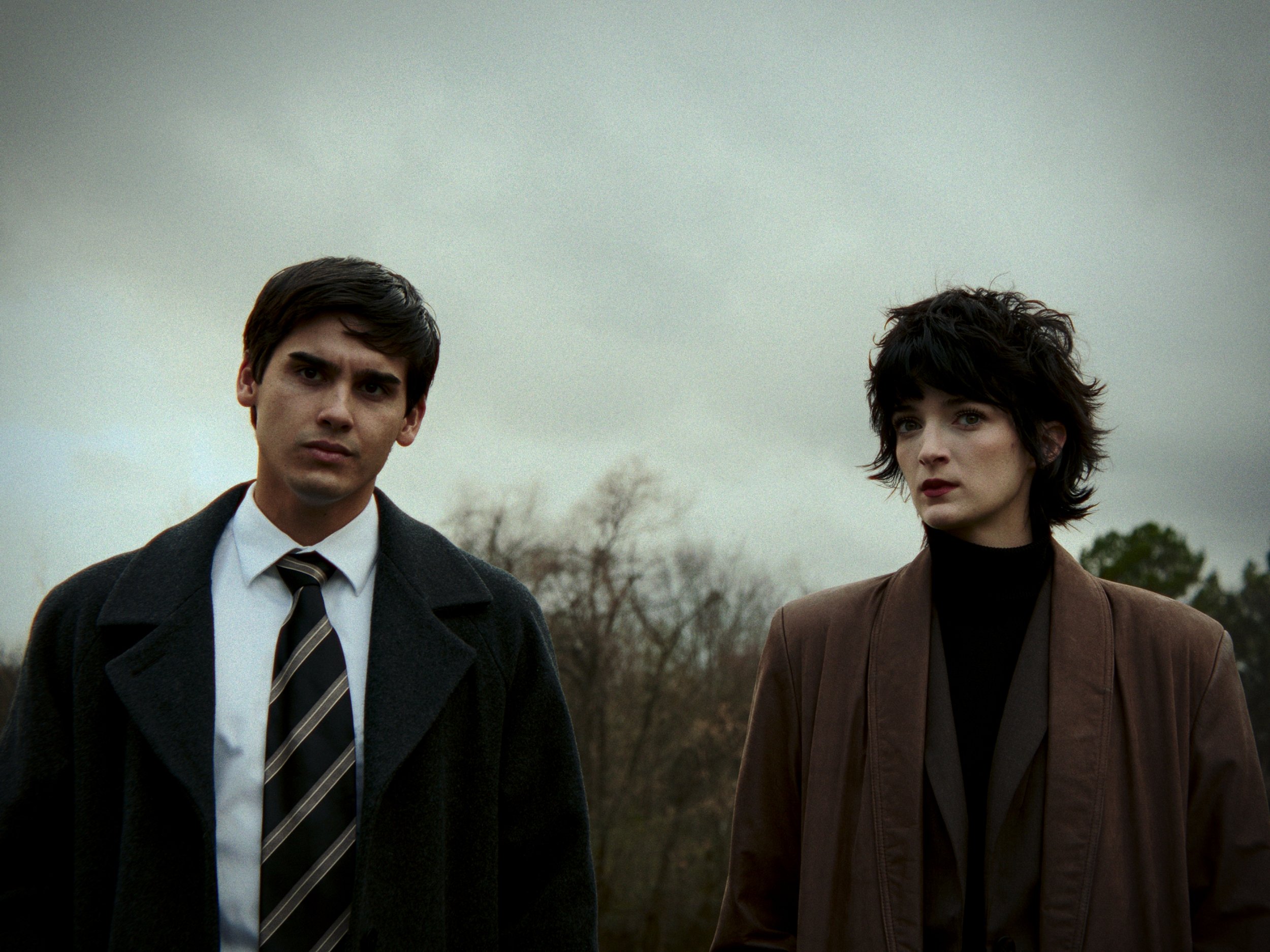 A young man and woman standing outdoors on a cloudy day, dressed in dark, stylish clothing, with serious expressions. X files vibes. Stranger Things. Joyce Chance