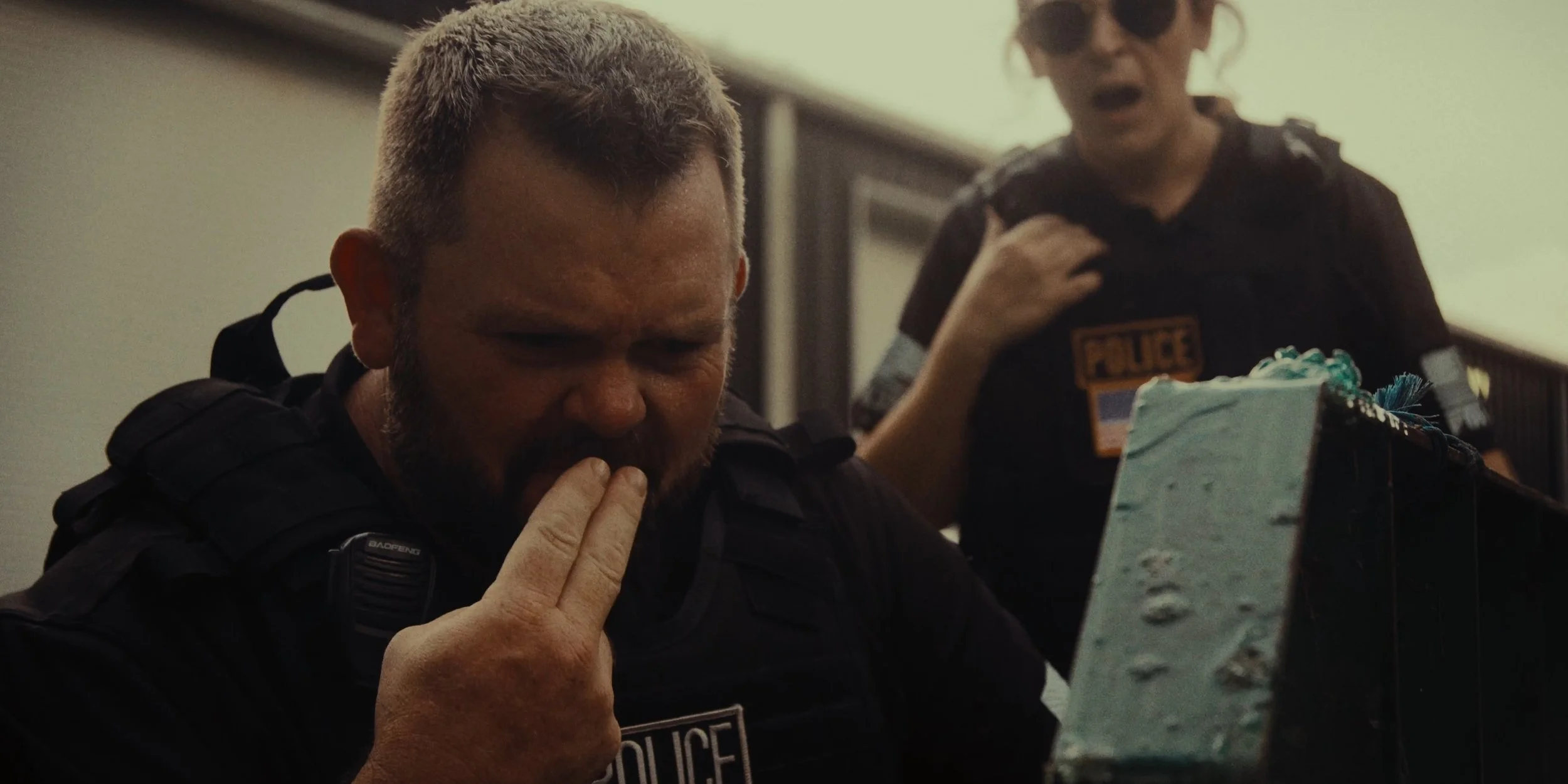Two police officers are in a room, one is contemplative with his hand near his mouth, and the other is talking.