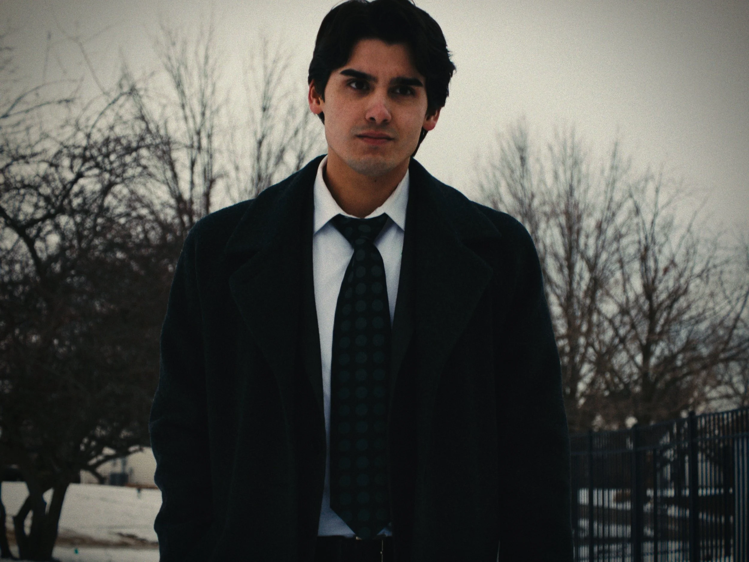 A young man with dark hair, wearing a black coat, white shirt, and dark polka dot tie, standing outdoors on a cloudy day with leafless trees and a metal fence in the background.