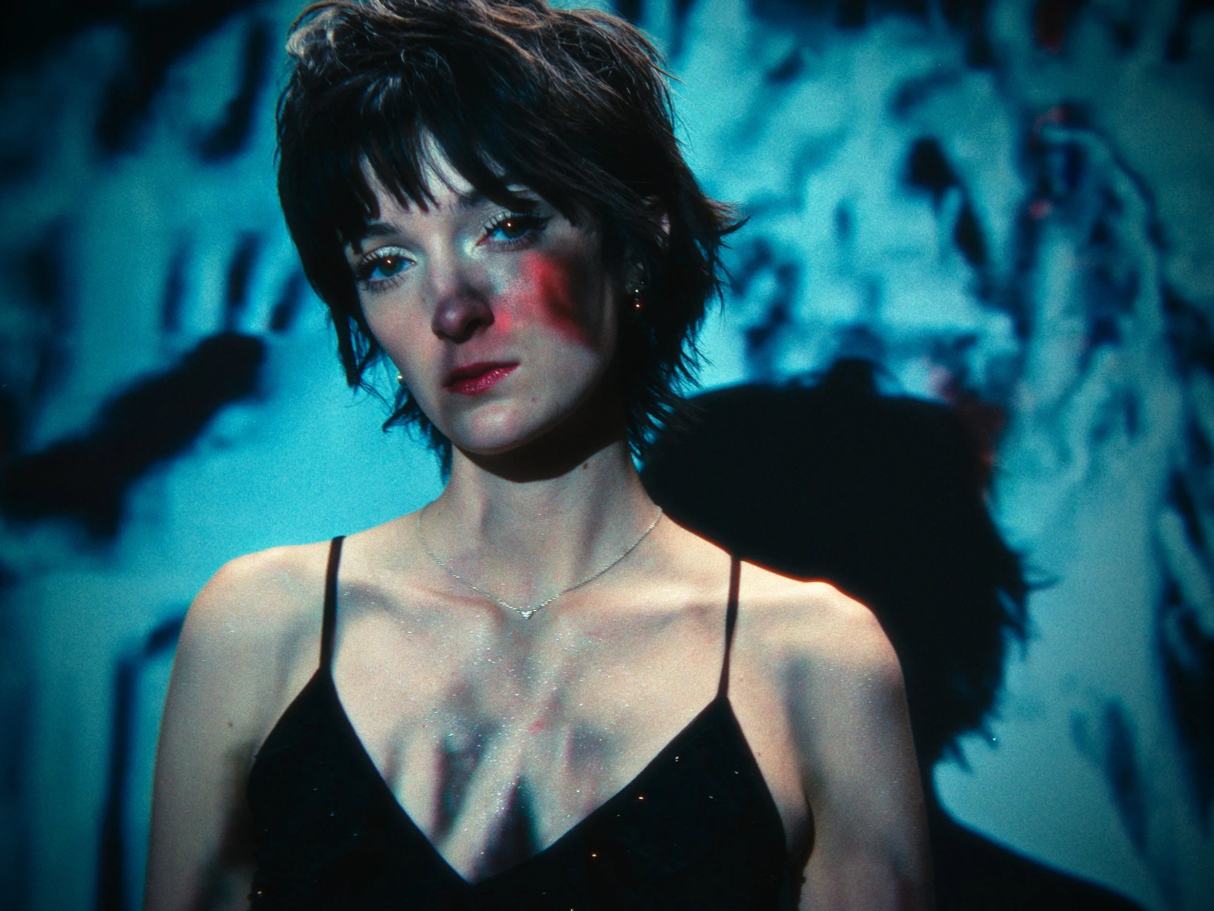 A woman with short dark hair, wearing a black spaghetti strap dress, stands in front of a wall with graffiti, with shadows falling across her face and chest.