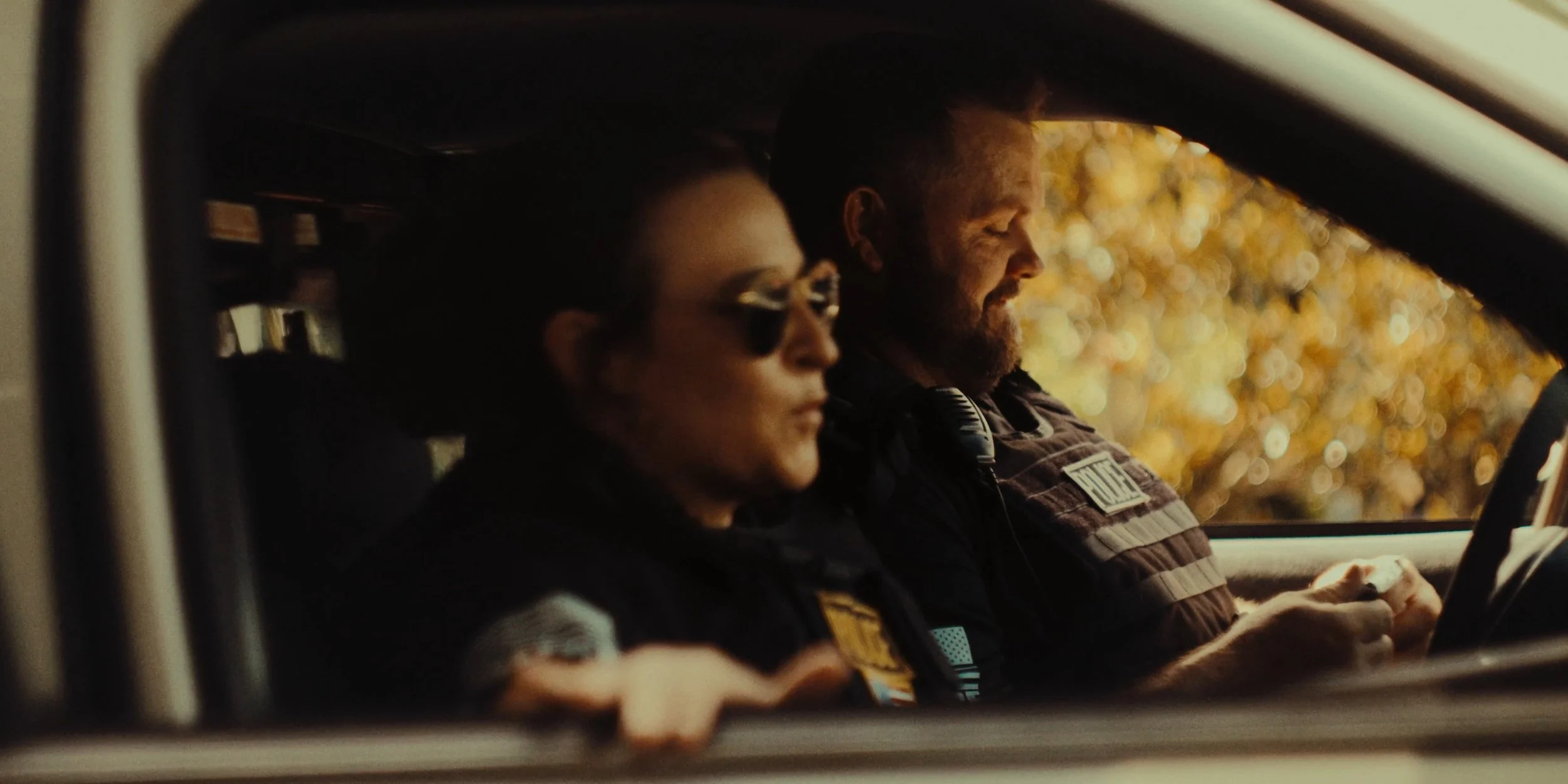 Two police officers sitting in a vehicle, focused, with autumn foliage visible outside the window.