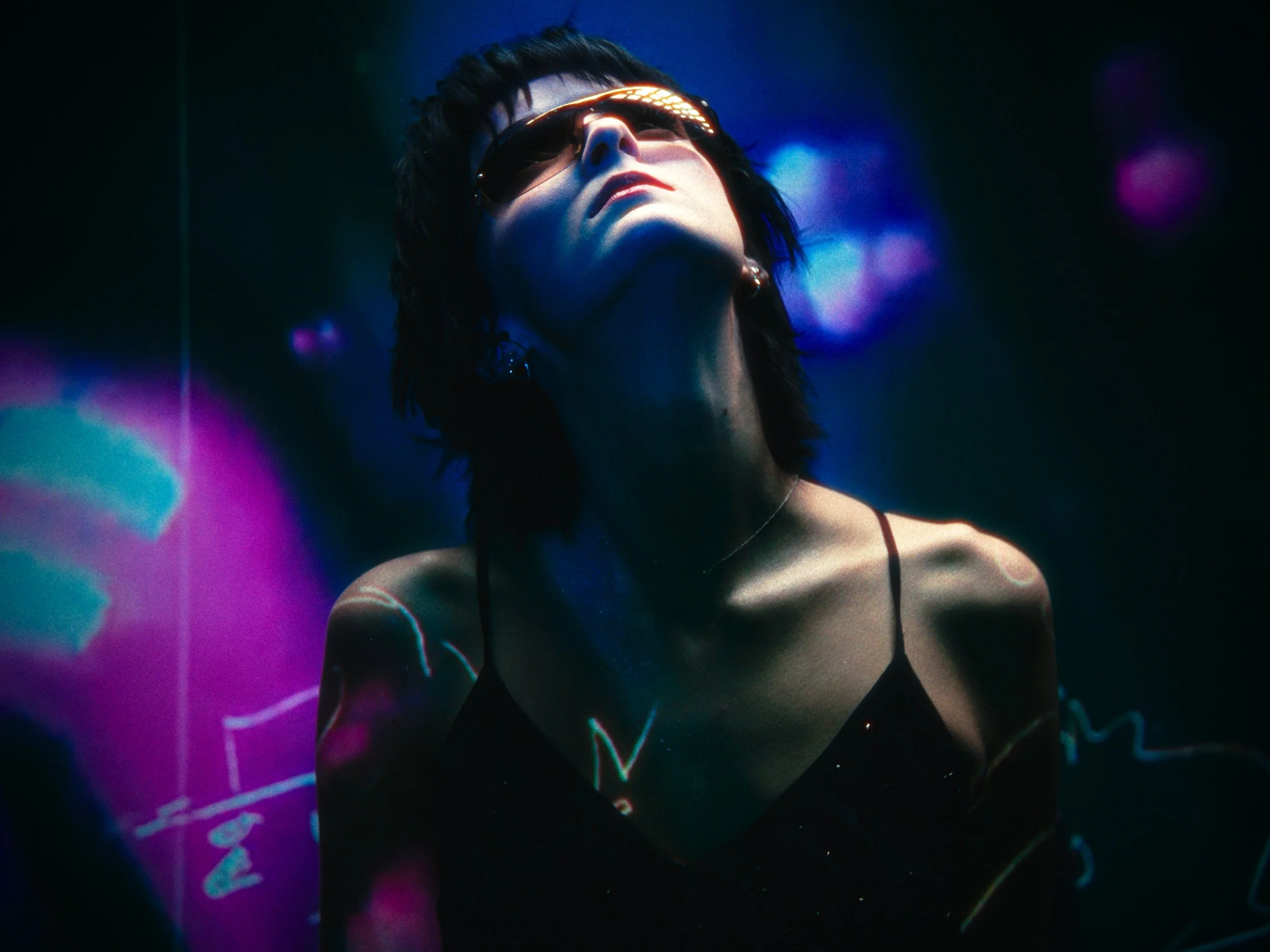 A woman with short dark hair wearing sunglasses and a black spaghetti strap top, illuminated by colorful neon lights, in a dark setting.