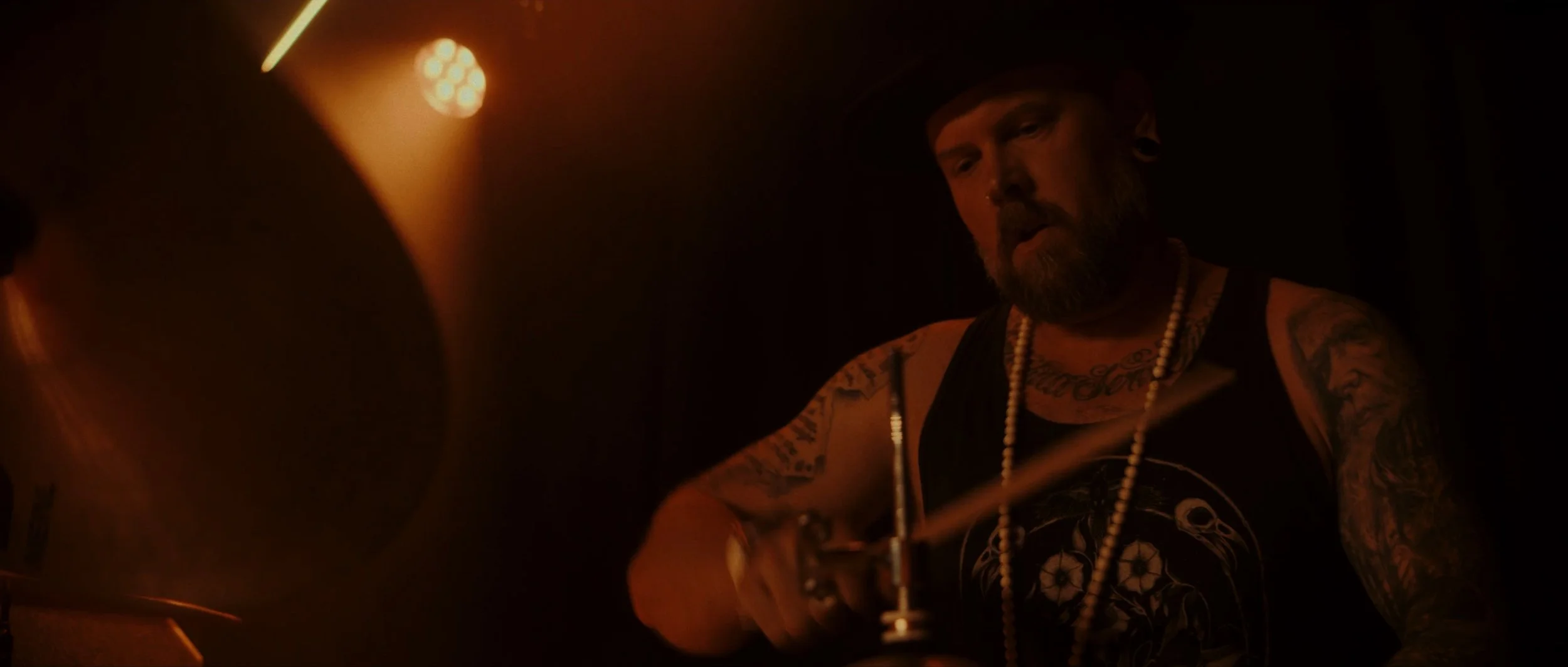 A bearded man with tattoos on his arms and chest, wearing a black tank top and a string of pearls, playing drums in a dimly lit, smoky environment.