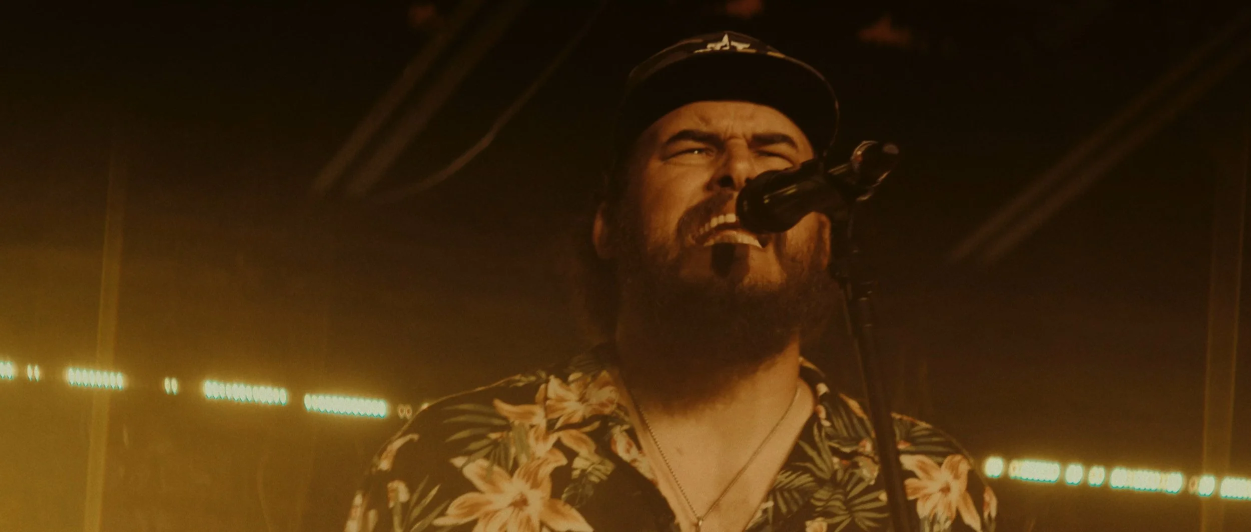A man singing passionately into a microphone on stage, wearing a floral shirt and a baseball cap, with stage lights in the background.