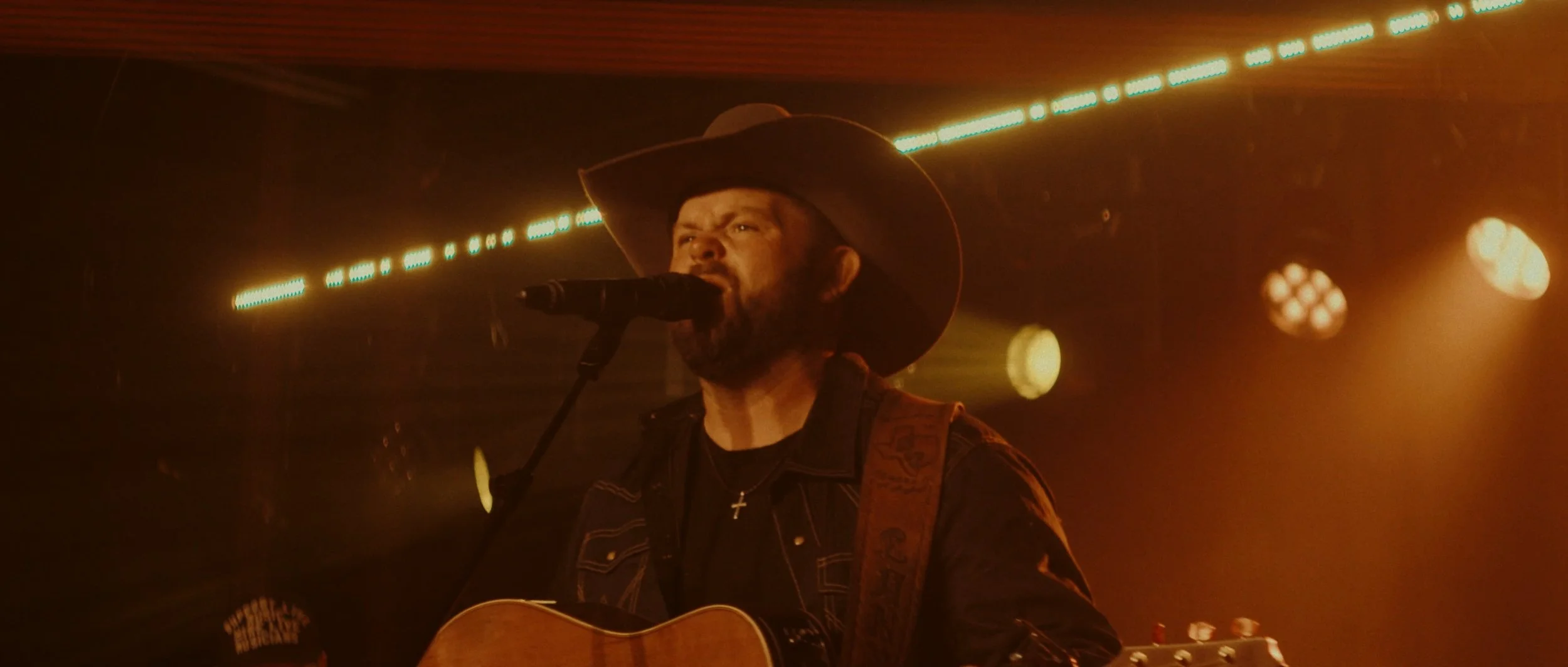 A man wearing a wide-brimmed hat and a necklace with a cross, singing into a microphone while playing an acoustic guitar on stage, illuminated by warm stage lighting.