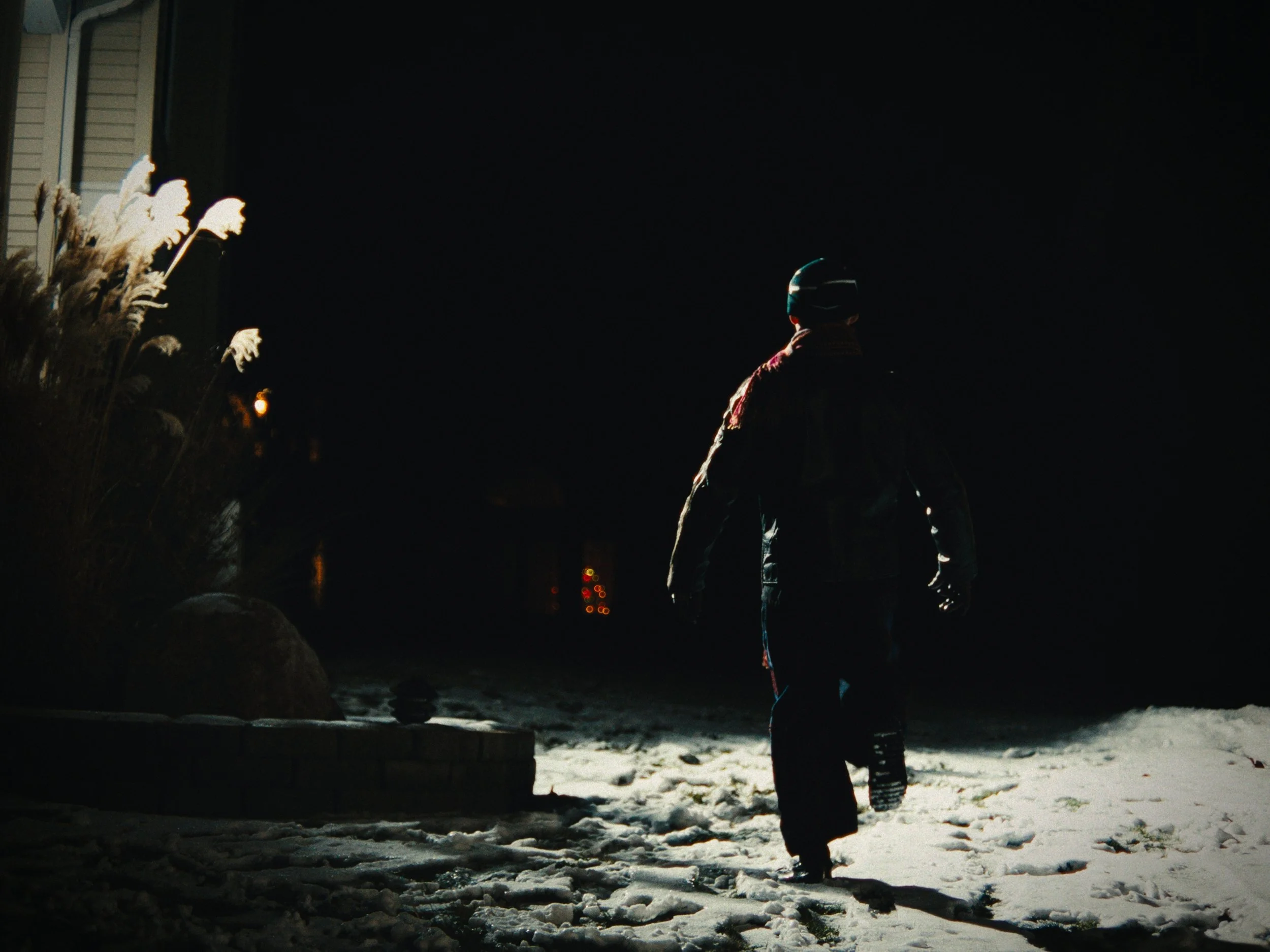 Person walking on snow-covered ground at night, illuminated by a light source behind and to the side, with a dark background and some house lights in the distance.