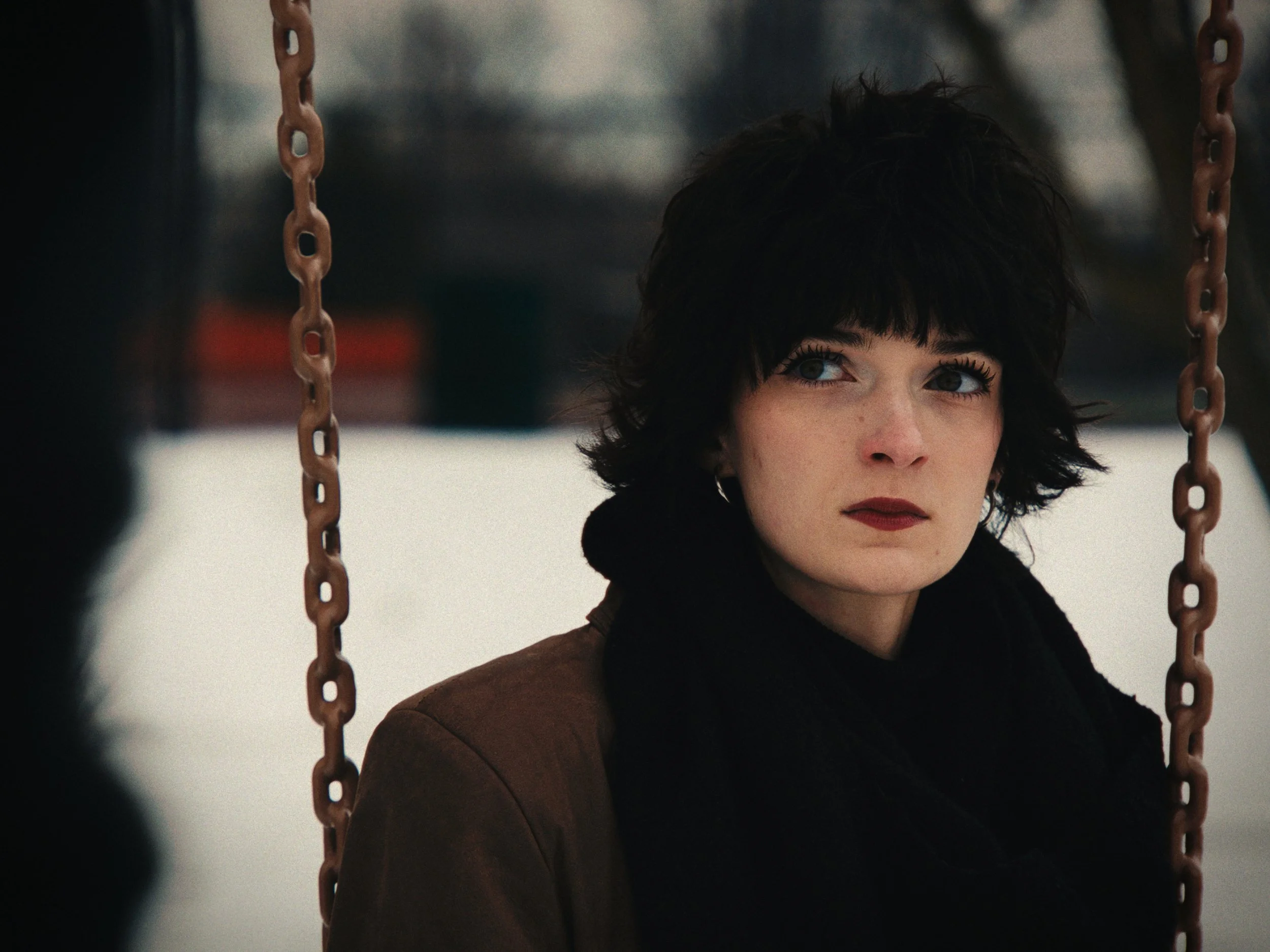 A woman with short dark hair and blue eyes sitting on a swing with chains, outdoors in winter.