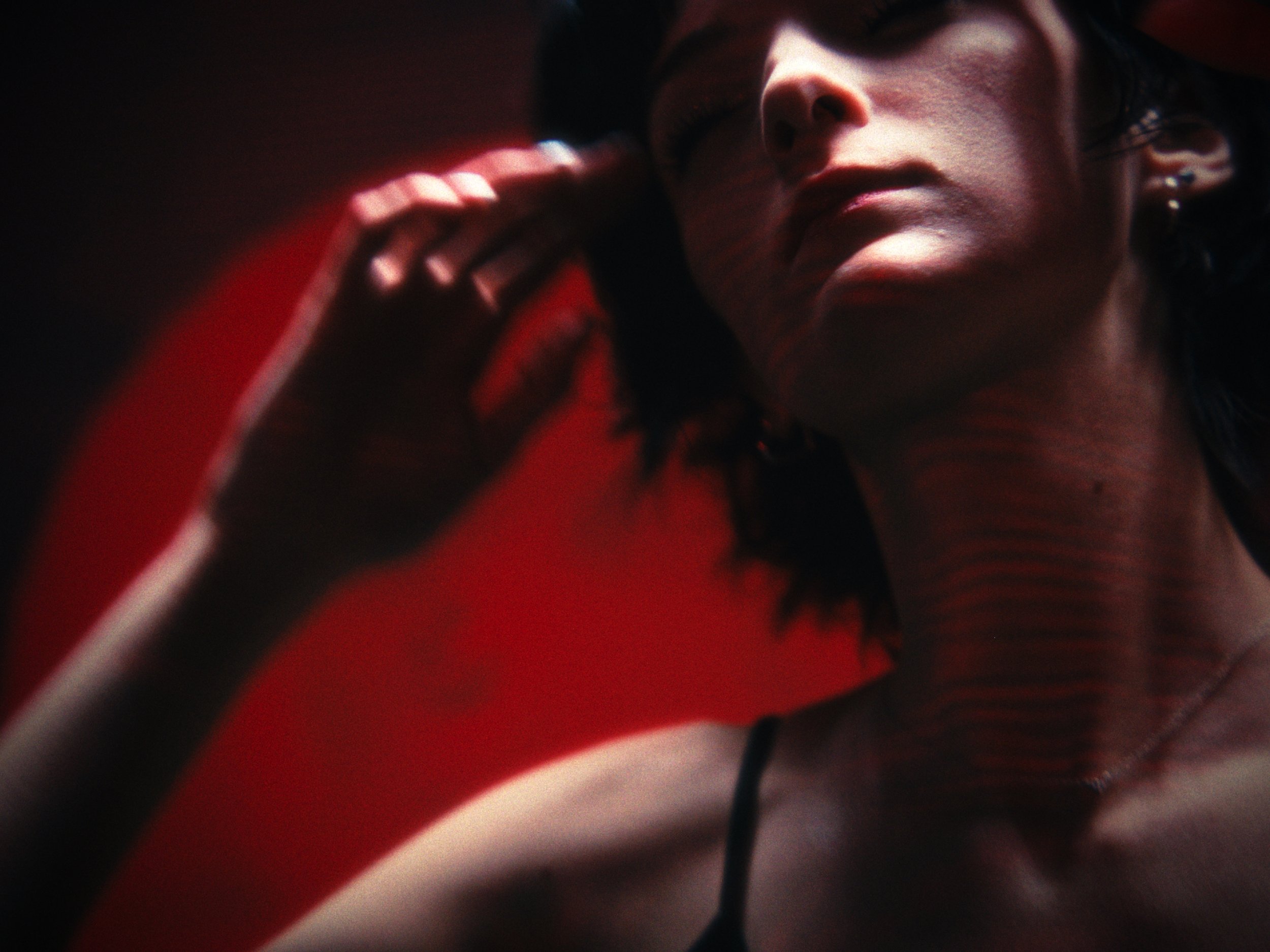 Close-up of a woman with short dark hair, wearing a black spaghetti strap top, touching her hair, with a red background.