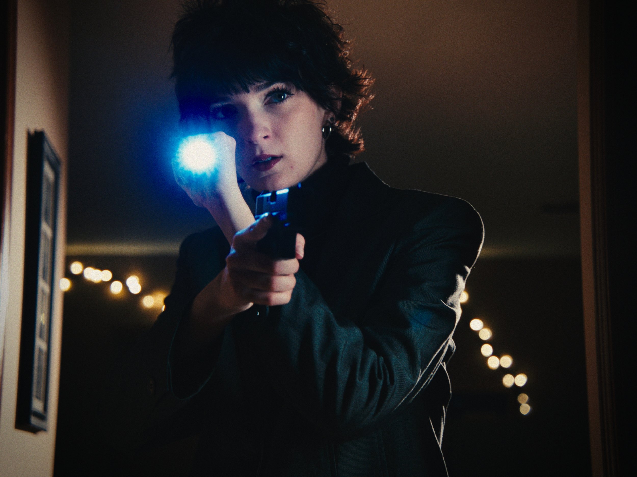 A woman with short dark hair, dressed in black, aiming a gun with a blue flashlight attached, in a dimly lit room with bokeh lights in the background.